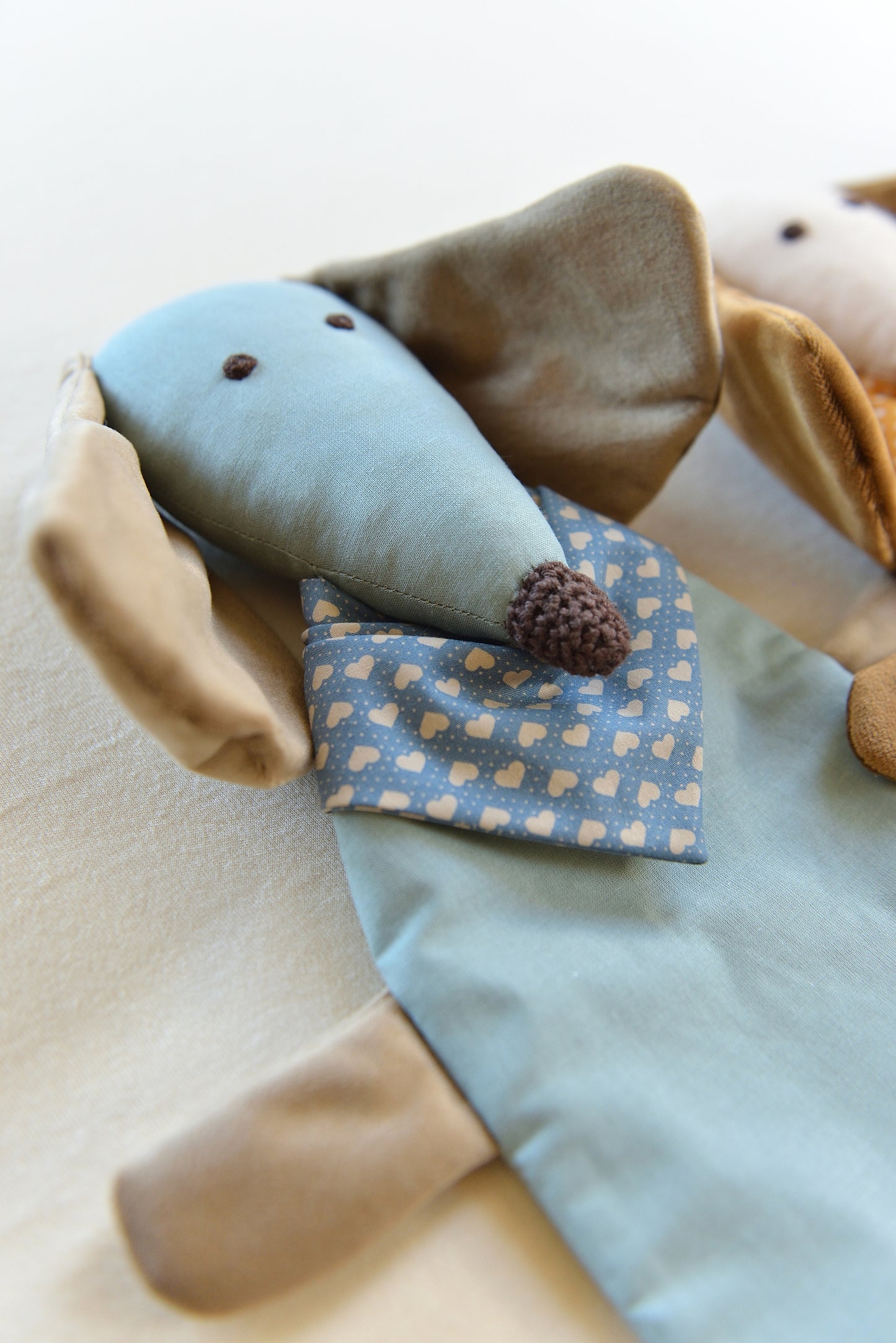 Dachshund Comforter shown from the side with long ears and snouts featuring a sky blue body and sand coloured shiny ears. It wears a heart patterned bandana in darker blue, conveying a playful and cozy feel.