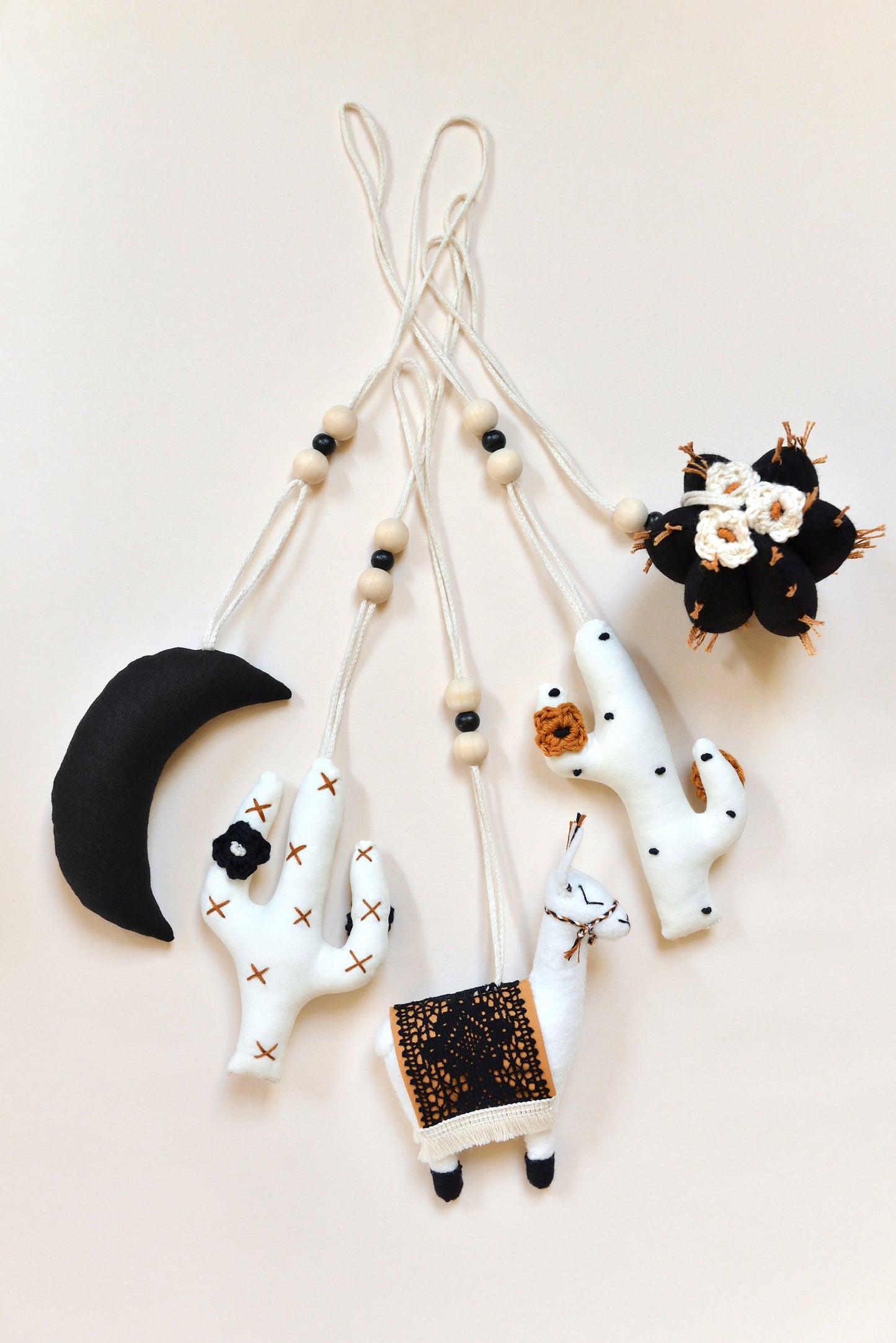 Flatlay featuring five handmade boho baby play gym toys on an ivory background: a black crescent moon, two white cacti with rust and black spikes and crocheted flowers, a llama with a rust saddle embellished with vintage style black lace, and a round black barell cactuc with rust spikes and ivory flowers. All hanging of strong macrame rope embellished with 3 wooden beads in the middle.