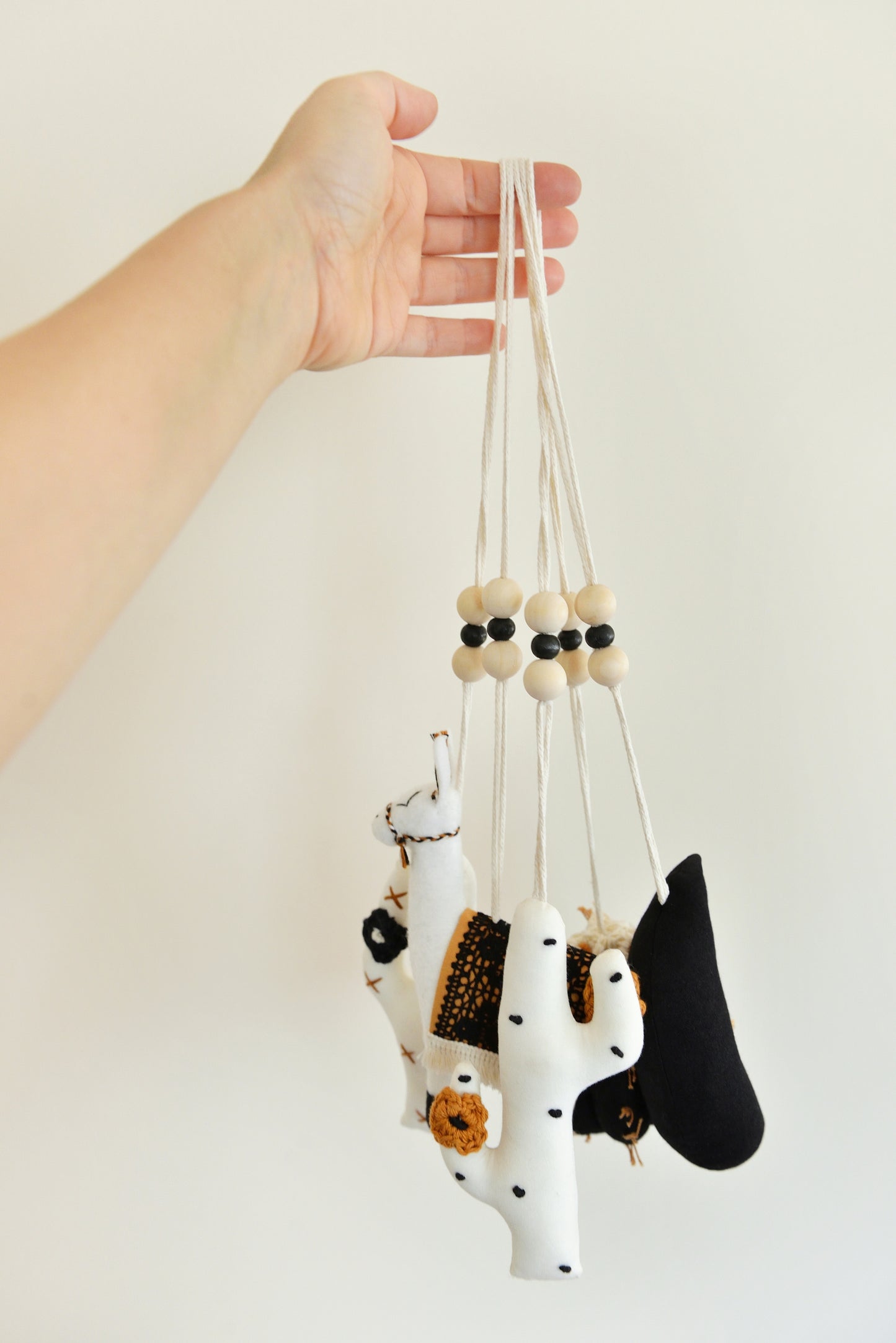 A hand holds a group of handmade boho play gym toys: a black barell cactus play gym toy with rust spikes and ivory flowers, a black crescent moon, two white cacti with rust and black spikes and crocheted flowers and a llama with a rust saddle embellished with vintage style black lace. All hanging of strong macrame rope embellished with 3 wooden beads in the middle.