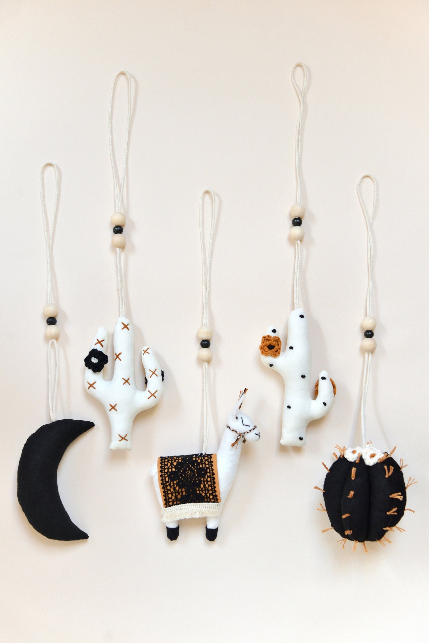 Flatlay featuring five handmade boho baby play gym toys on an ivory background: a black crescent moon, two white cacti with rust and black spikes and crocheted flowers, a llama with a rust saddle embellished with vintage style black lace, and a round black barell cactuc with rust spikes and ivory flowers. All hanging of strong macrame rope embellished with 3 wooden beads in the middle.