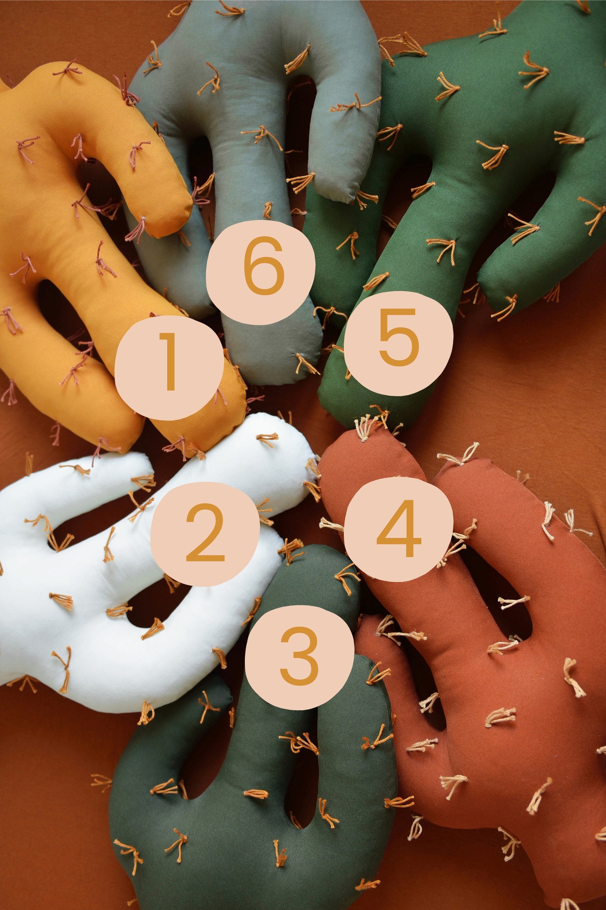Flatlay of saguaro shaped plush cactus pillows  in various colors—dark olive, marigold, offwhite, khaki, rust, juniper green—placed on a mattress with rust fitted sheet. Numbers on pillows from 1-6 help choose style.