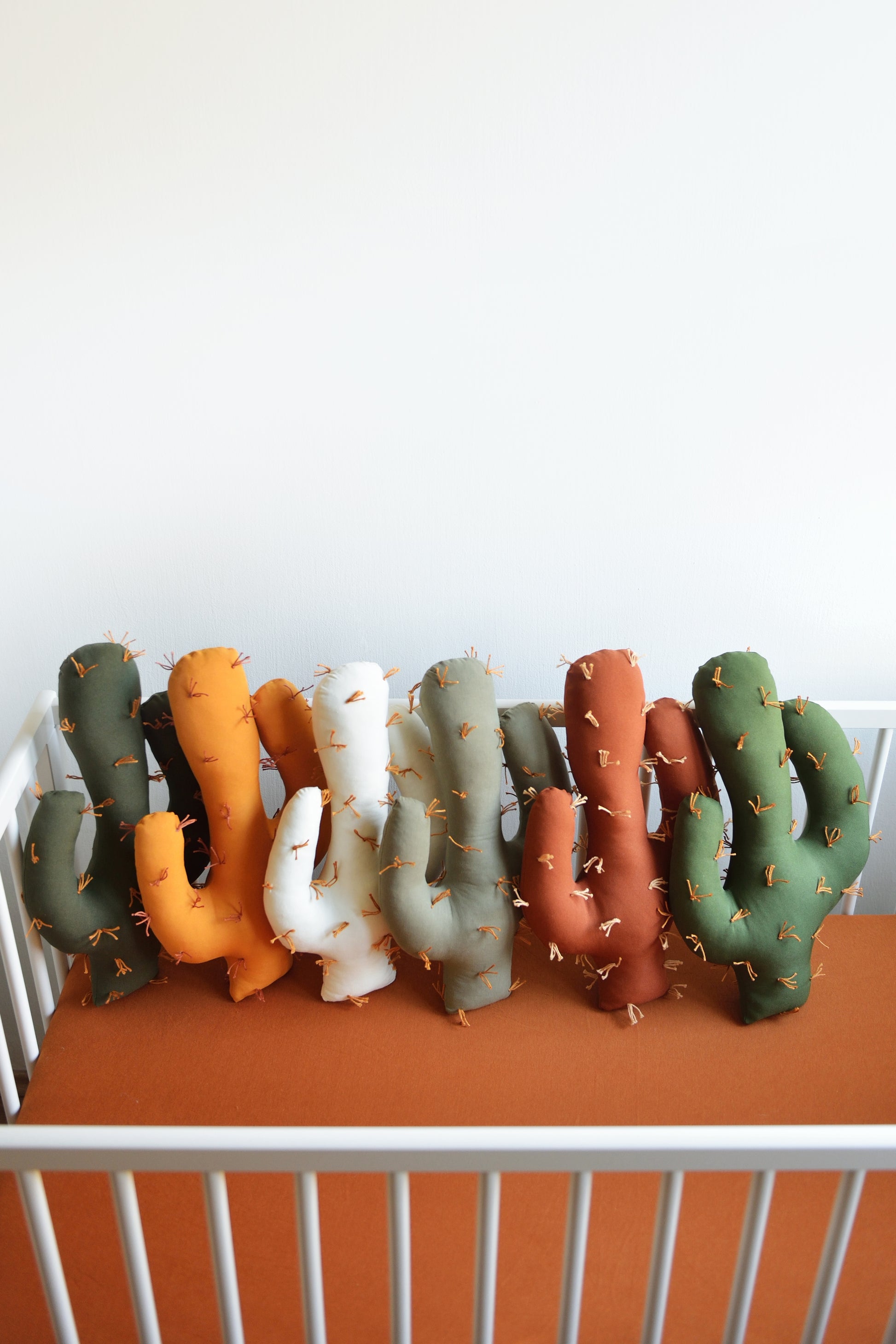 Saguaro shaped plush cactus pillows  in various colors—dark olive, marigold, offwhite, khaki, rust, juniper green—sit in a row in a white crib, on a mattress with rust fitted sheet.