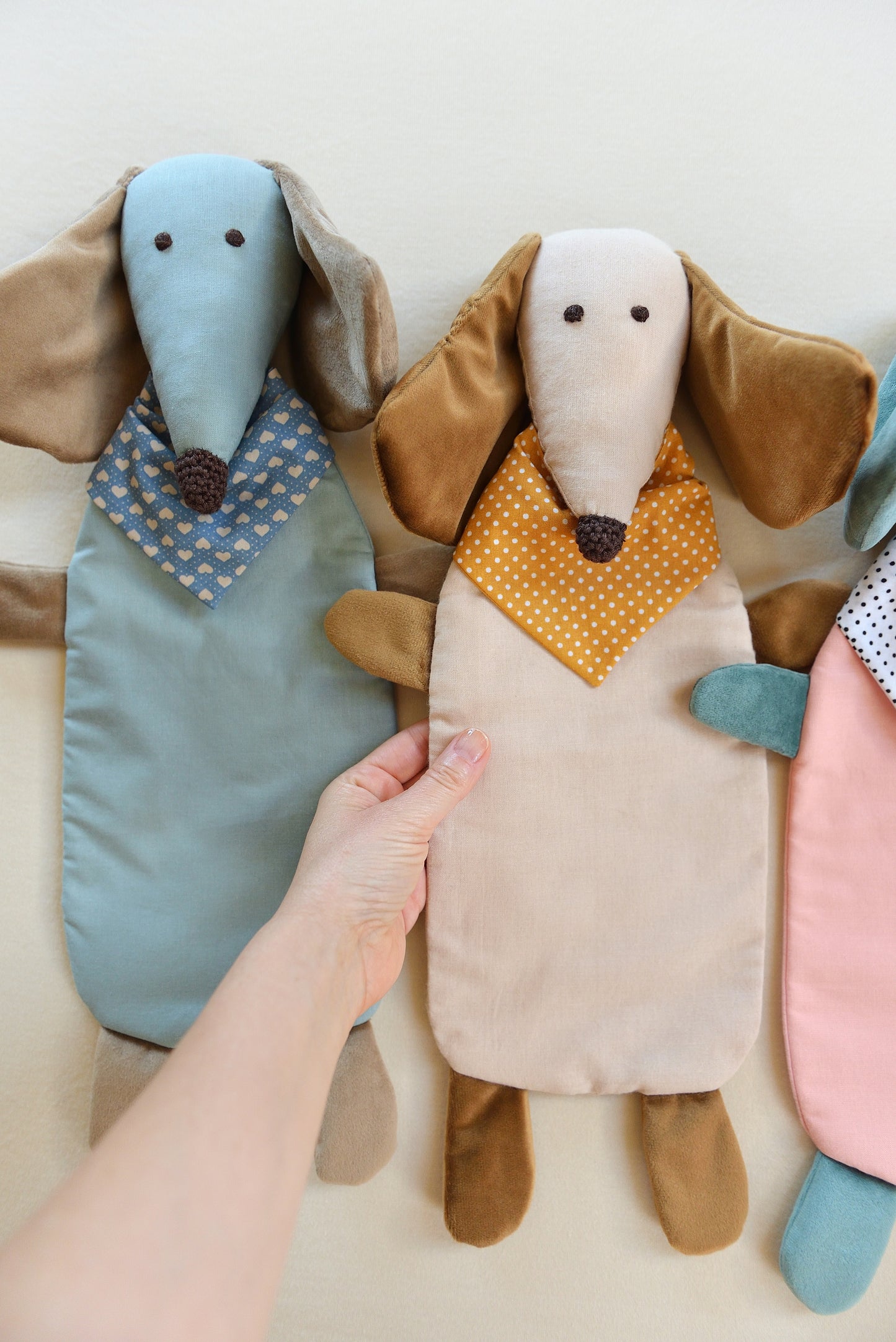 Left hand holding one of three Dachshund Comforters with long ears and snouts in blue, cream, and pink. Each wears a patterned bandana.