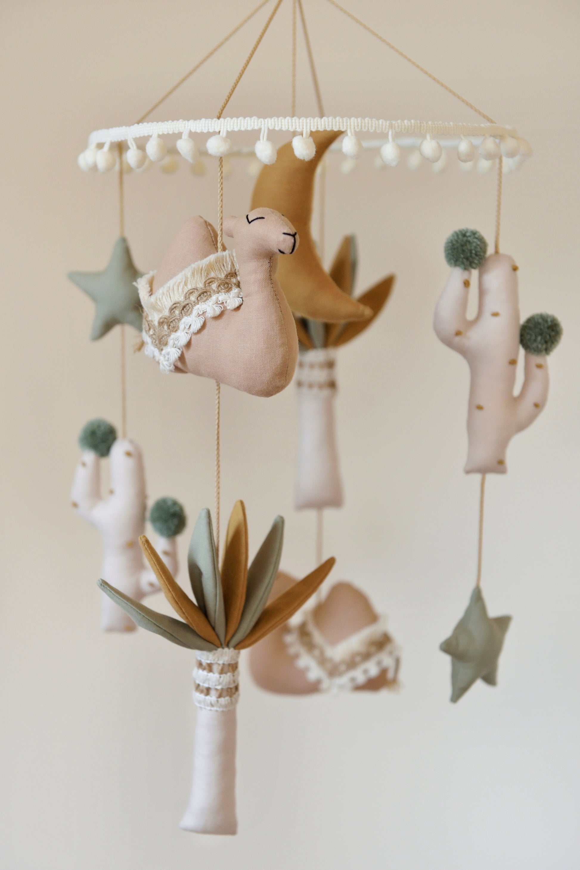 Desert-themed mobile with tan camels, sage stars, ivory and sage palm trees, cacti and a gold mustard moon suspended from a hoop adorned with ivory pom pom trim.