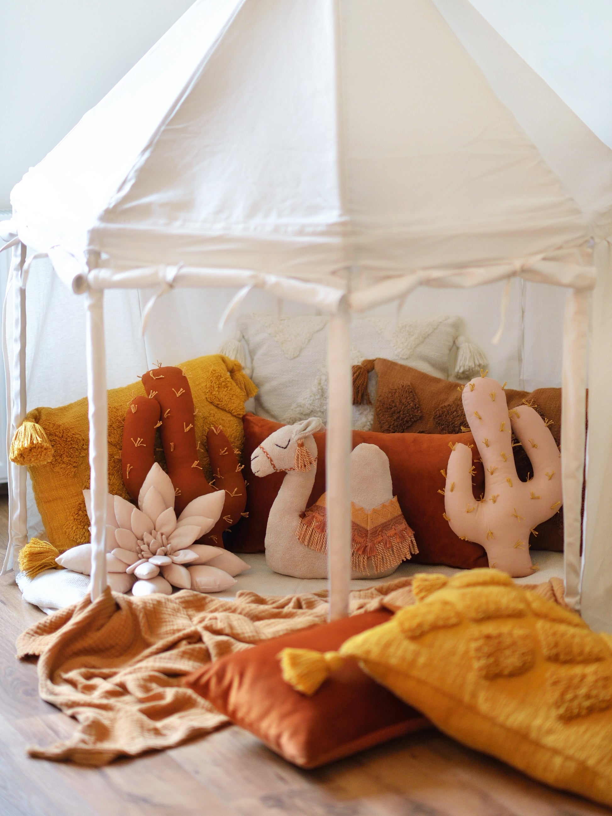 A cozy ivory play tent filled with cactus-shaped pillows and a camel plush toy. Warm rust and ochre tones create a comforting, playful atmosphere.