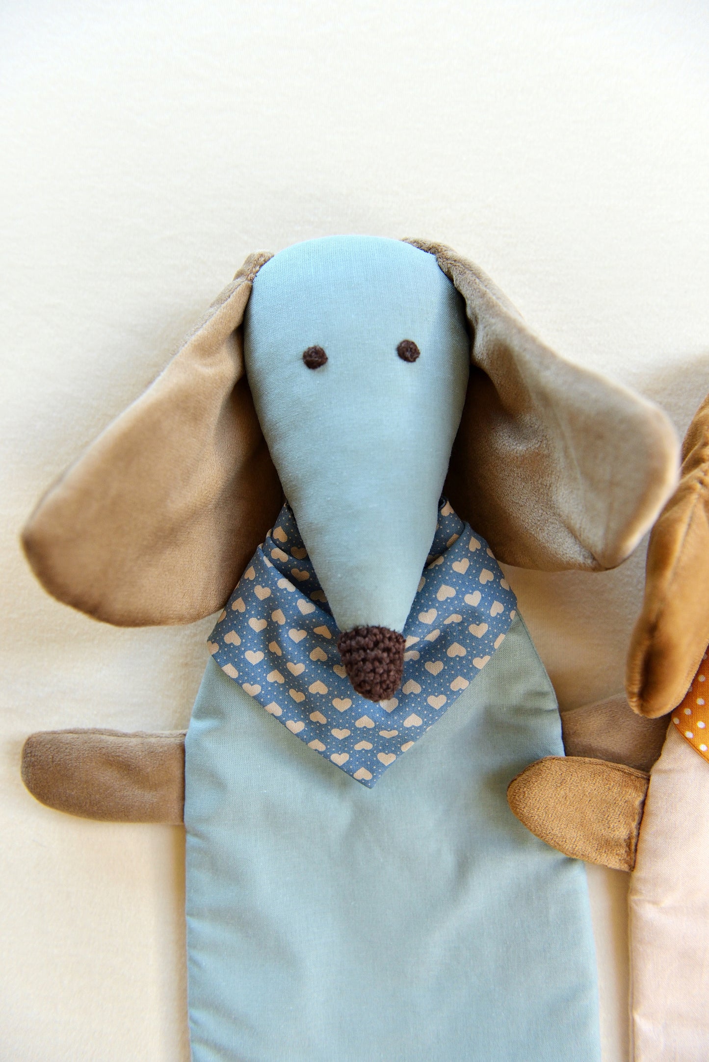 Dachshund Comforter up close with long ears and snouts featuring a sky blue body and sand coloured shiny ears. It wears a heart patterned bandana in darker blue, conveying a playful and cozy feel.