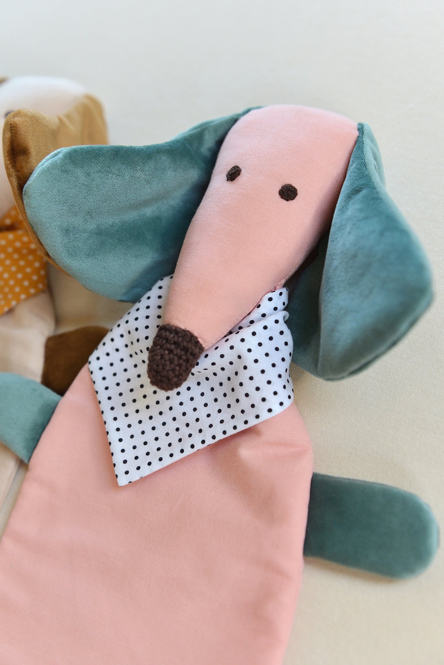 Dachshund Comforter from the side with long ears and snouts featuring a pink body and teal coloured shiny ears. It wears a white bandana with black dots conveying a playful and cozy feel.