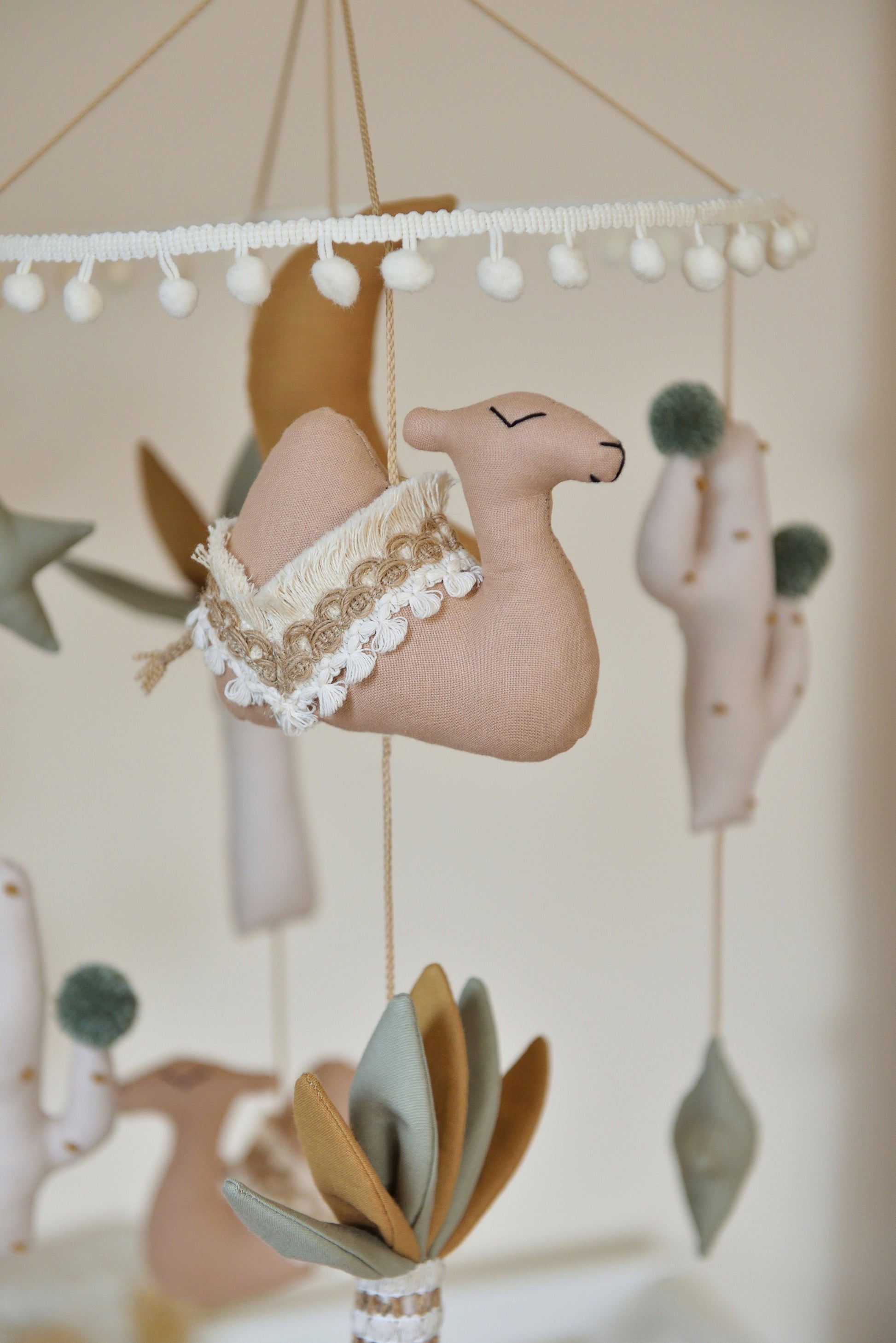 Close up of a desert-themed mobile with tan camels, sage stars, ivory and sage palm trees, cacti and a gold mustard moon in the middle hangs above a white crib.