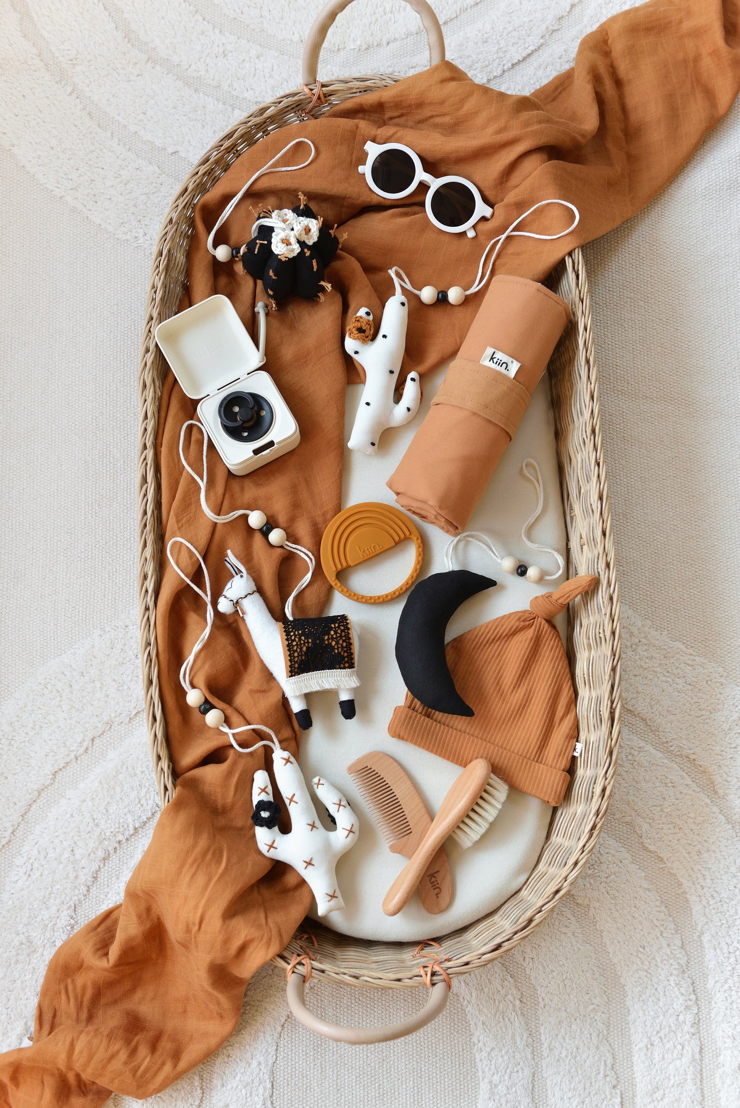 A wicker baby changing basket holds baby items placed on a rust-colored swaddle: sunglasses, five handmade llama, moon and cactus baby play gym toys, pacifier and storage box, cotton beanie, wooden comb set, travel change mat and silicone teether creating a cozy feel.