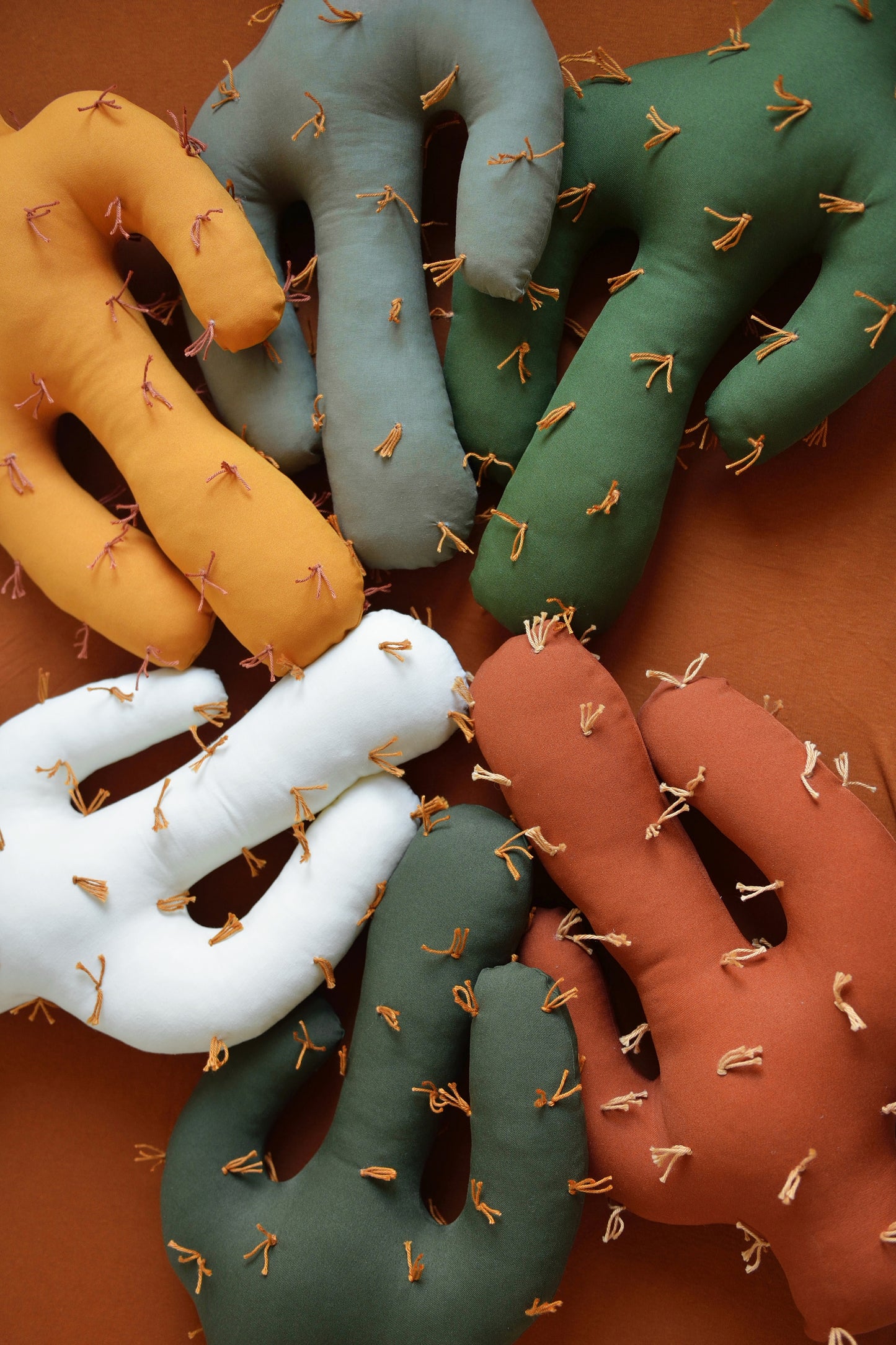 Flatlay of saguaro shaped plush cactus pillows  in various colors—dark olive, marigold, offwhite, khaki, rust, juniper green—placed on a mattress with rust fitted sheet.