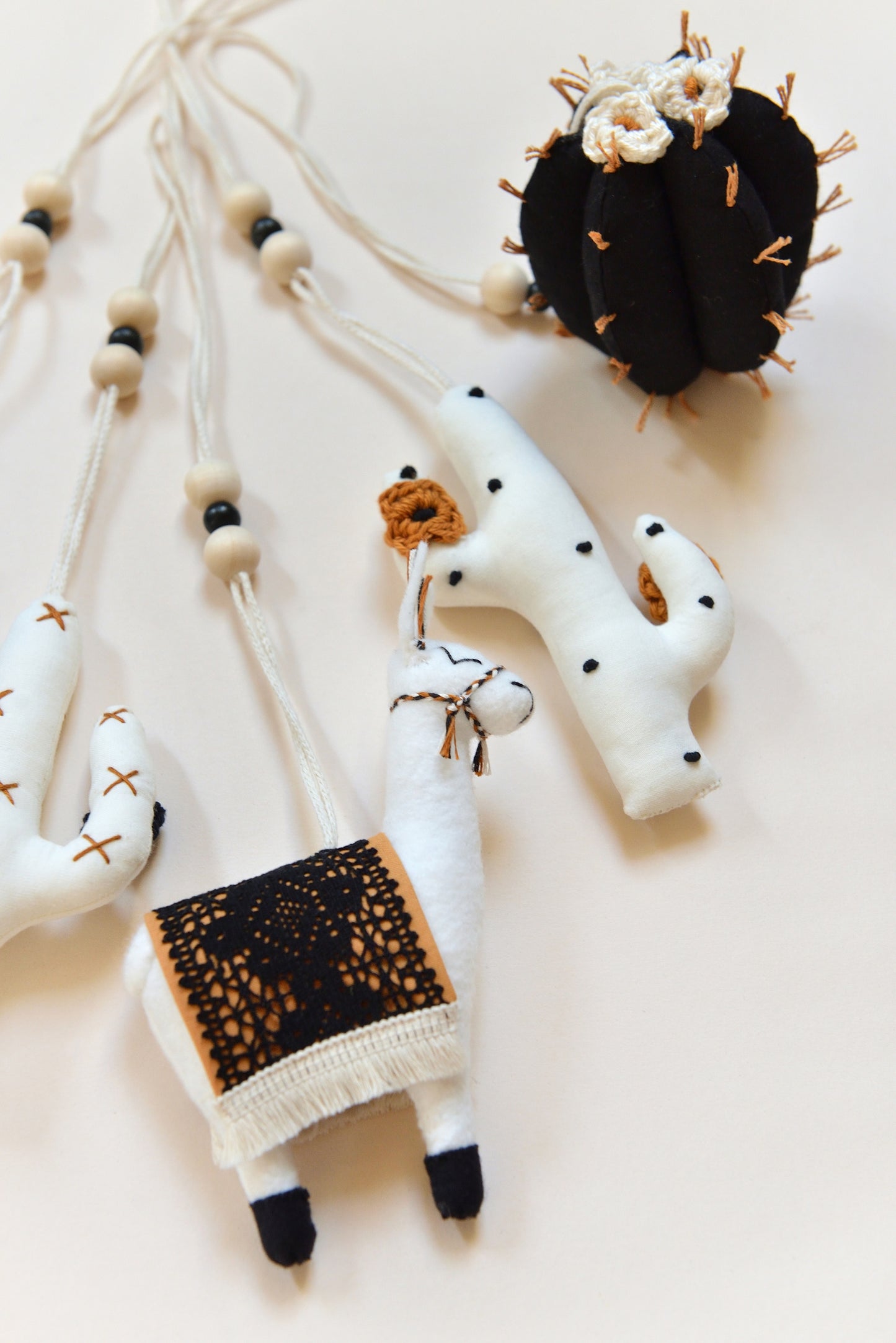 Close up of flatlay featuring four handmade boho baby play gym toys on an ivory background: two white cacti with rust and black spikes and crocheted flowers, a llama with a rust saddle embellished with vintage style black lace, and a round black barell cactuc with rust spikes and ivory flowers. All hanging of strong macrame rope embellished with 3 wooden beads in the middle.