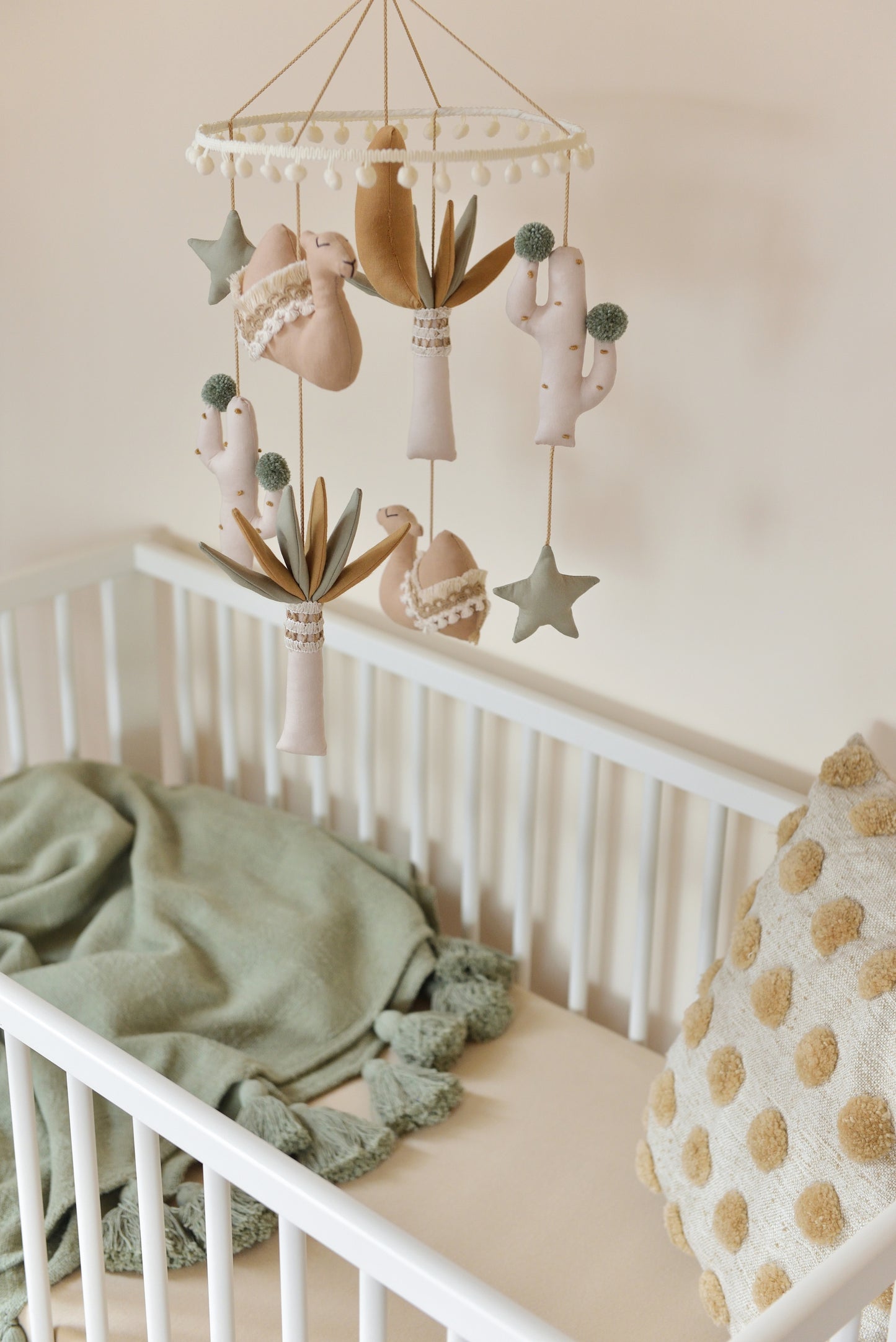 A cozy gender-neutral boho nursery with a white crib. A desert-themed mobile with tan camels, sage stars, ivory and sage palm trees and a gold mustard moon in the middle hangs above. On the right Throw Pillow in the crib, next to a sage blanket with tassels.