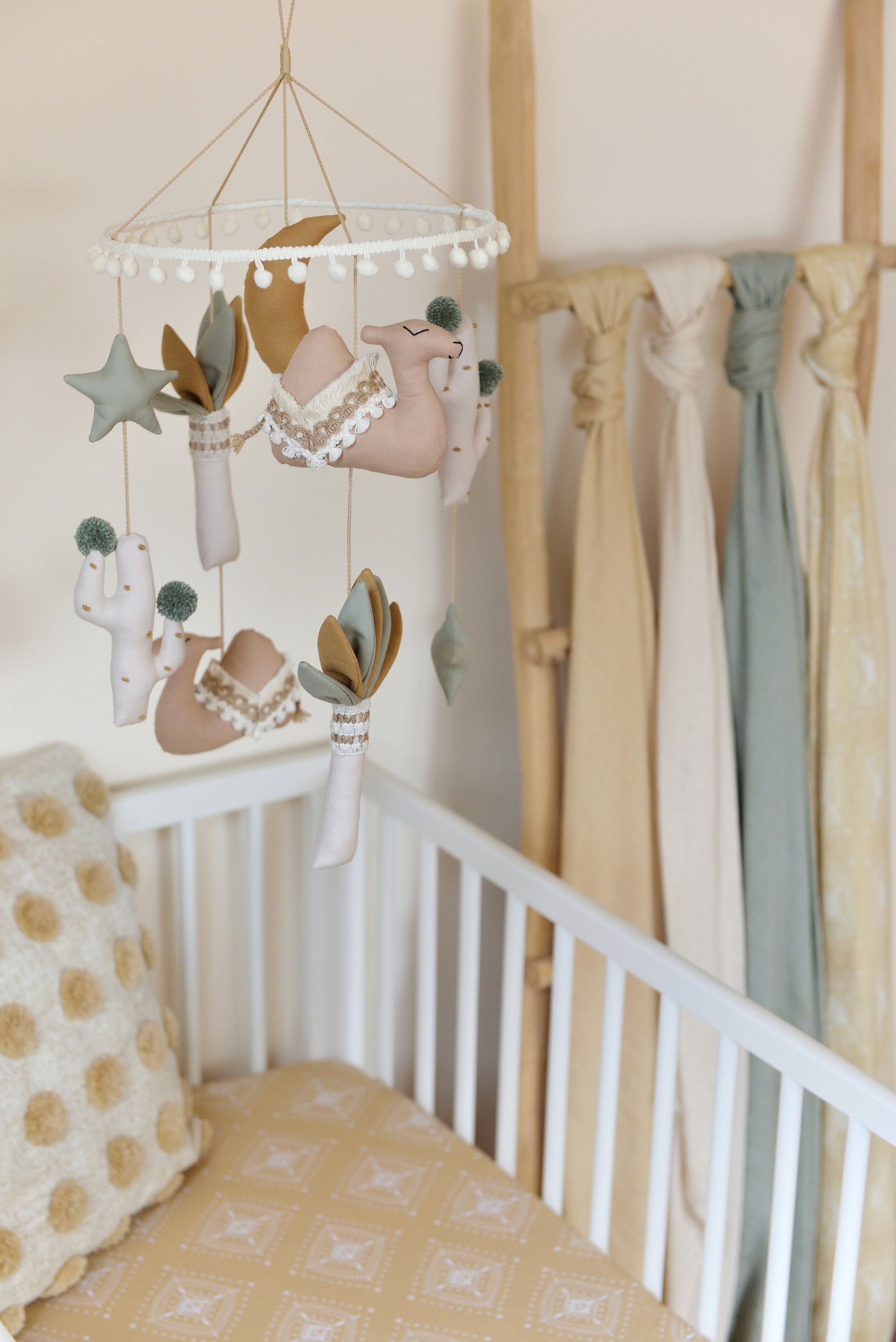 A cozy gender-neutral boho nursery with a white crib and wooden ladder holding neutral coloured swaddles in the background. A desert-themed mobile with tan camels, sage stars, ivory and sage palm trees and a gold mustard moon in the middle hangs above. Under a Throw Pillow in the crib on an oat colour patterned crib sheet.
