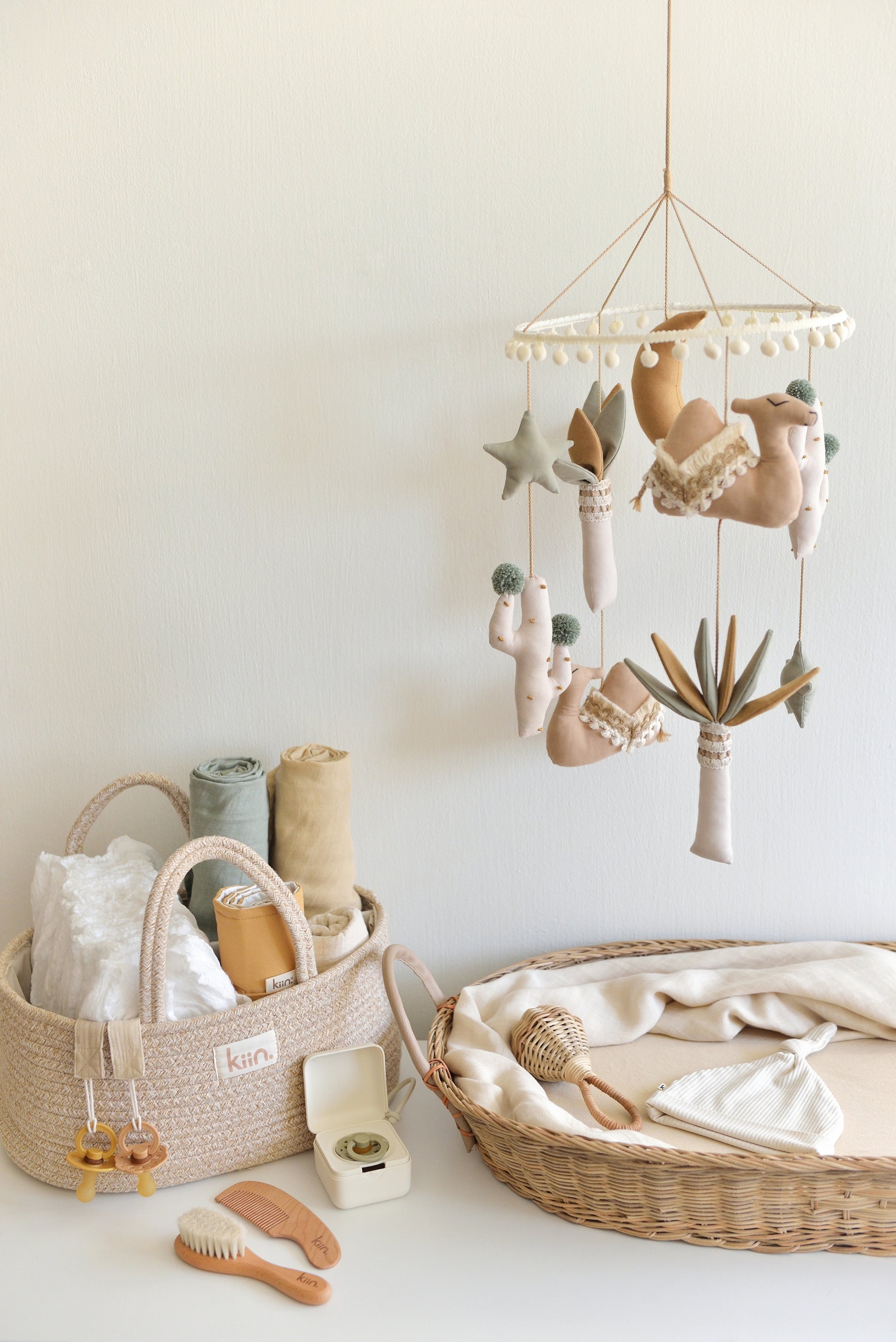 Gender-neutral boho nursery setup with a desert-themed camel mobile hanging over the changing table. Nappy caddy on the left, and a wicker changing basket with newborn essentials on the right.