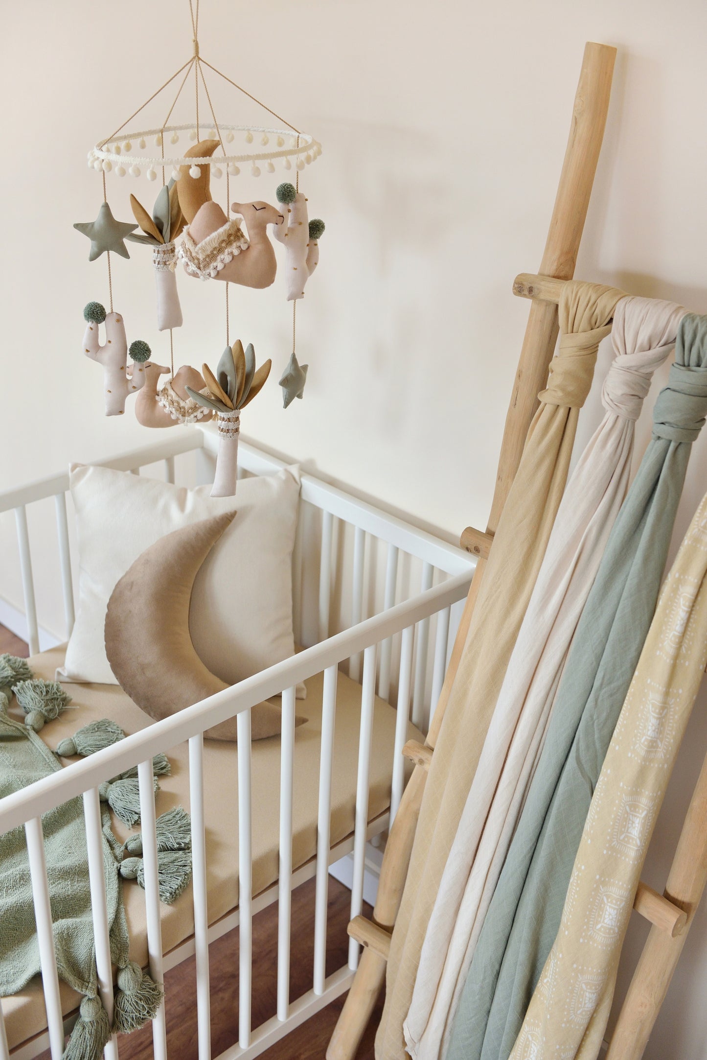 A cozy gender-neutral boho nursery with a white crib and wooden ladder holding neutral coloured swaddles. A desert-themed mobile with tan camels, sage stars, ivory and sage palm trees and a gold mustard moon in the middle hangs above. Under a Moon Decor Pillow and Ivory Throw Pillow in the crib, next to a sage blanket with tassels.