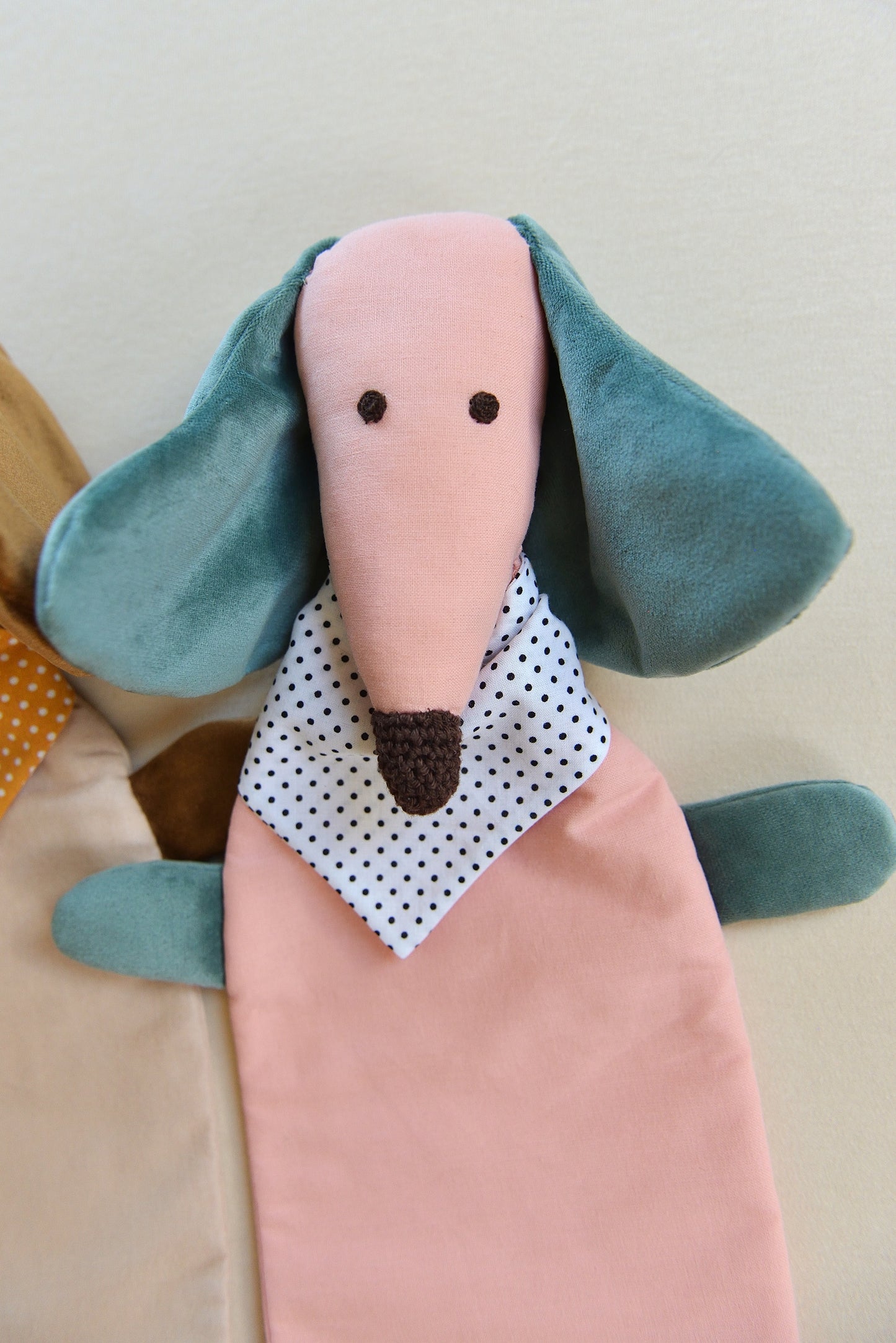 Dachshund Comforter up close with long ears and snouts featuring a pink body and teal coloured shiny ears. It wears a white bandana with black dots conveying a playful and cozy feel.