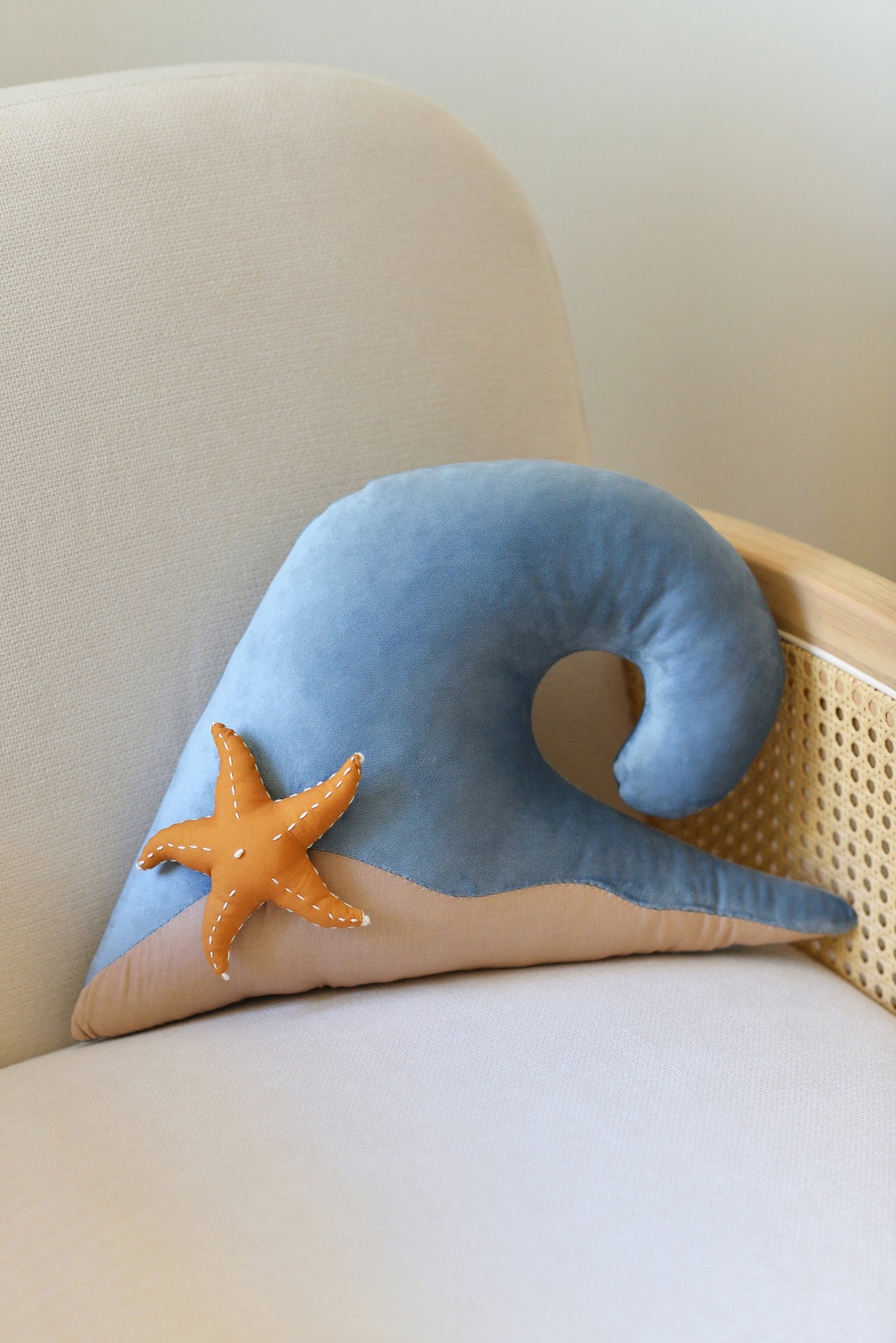 Ocean wave shaped decor pillow with a starfish placed on a rattan armchair against a white wall.