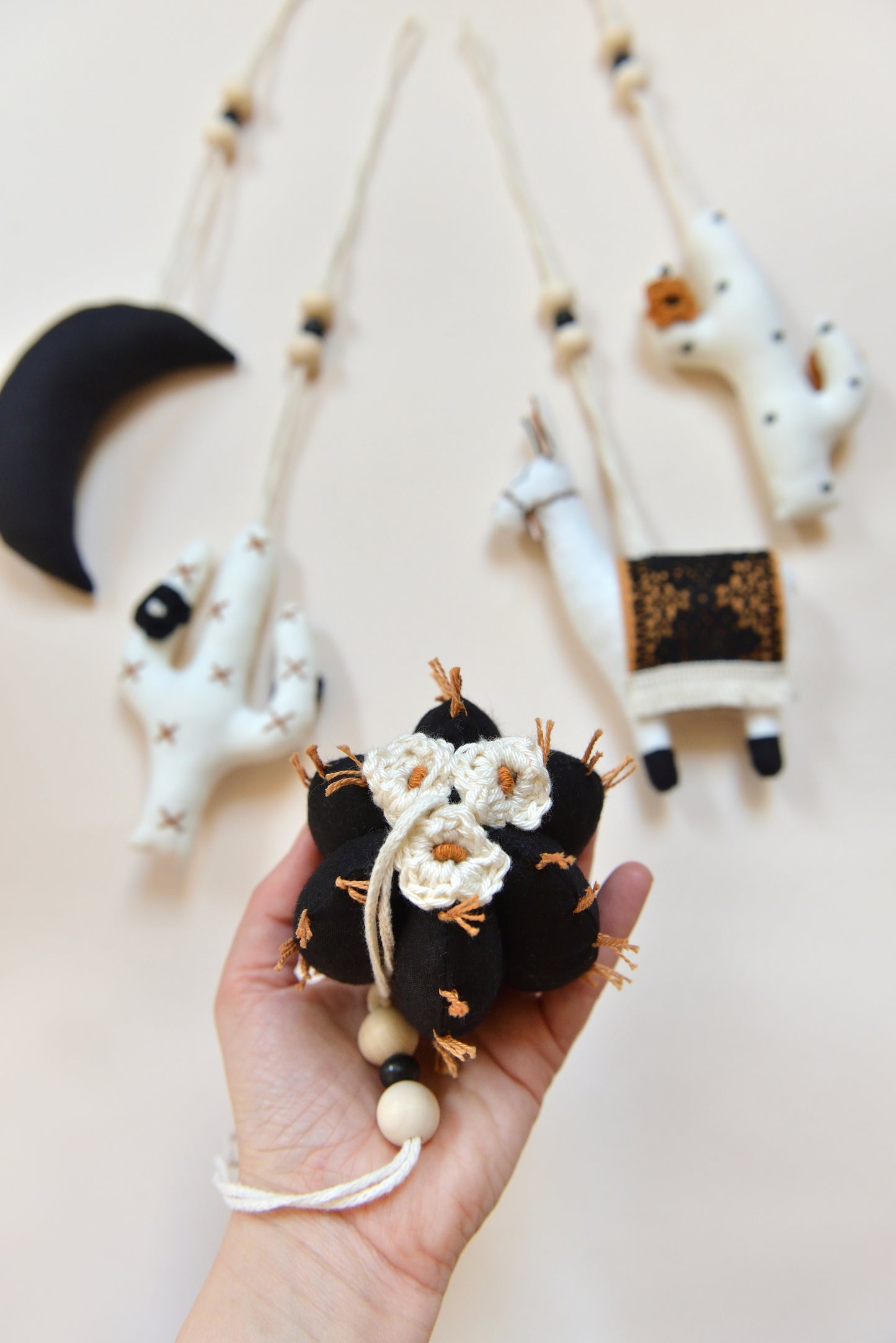 A hand holds a handmade black barell cactus play gym toy with rust spikes and ivory flowers, set against a flatlay of hanging play gym toys: a black crescent moon, two white cacti with rust and black spikes and crocheted flowers and a llama with a rust saddle embellished with vintage style black lace. All hanging of strong macrame rope embellished with 3 wooden beads in the middle.