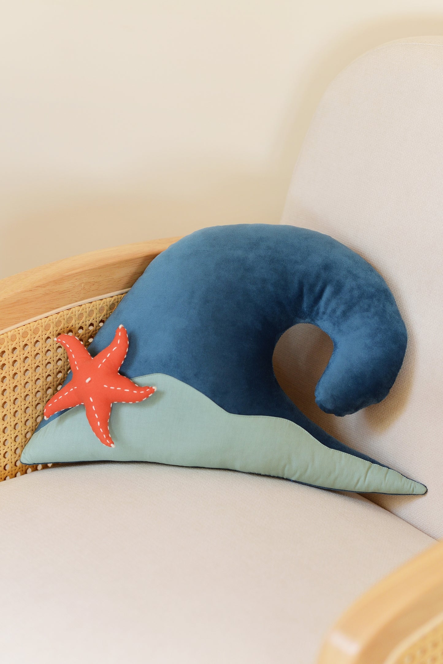 Ocean Blue wave shaped decor pillow with dark mint green sandy base and coral red starfish on an ivory rattan armchair.