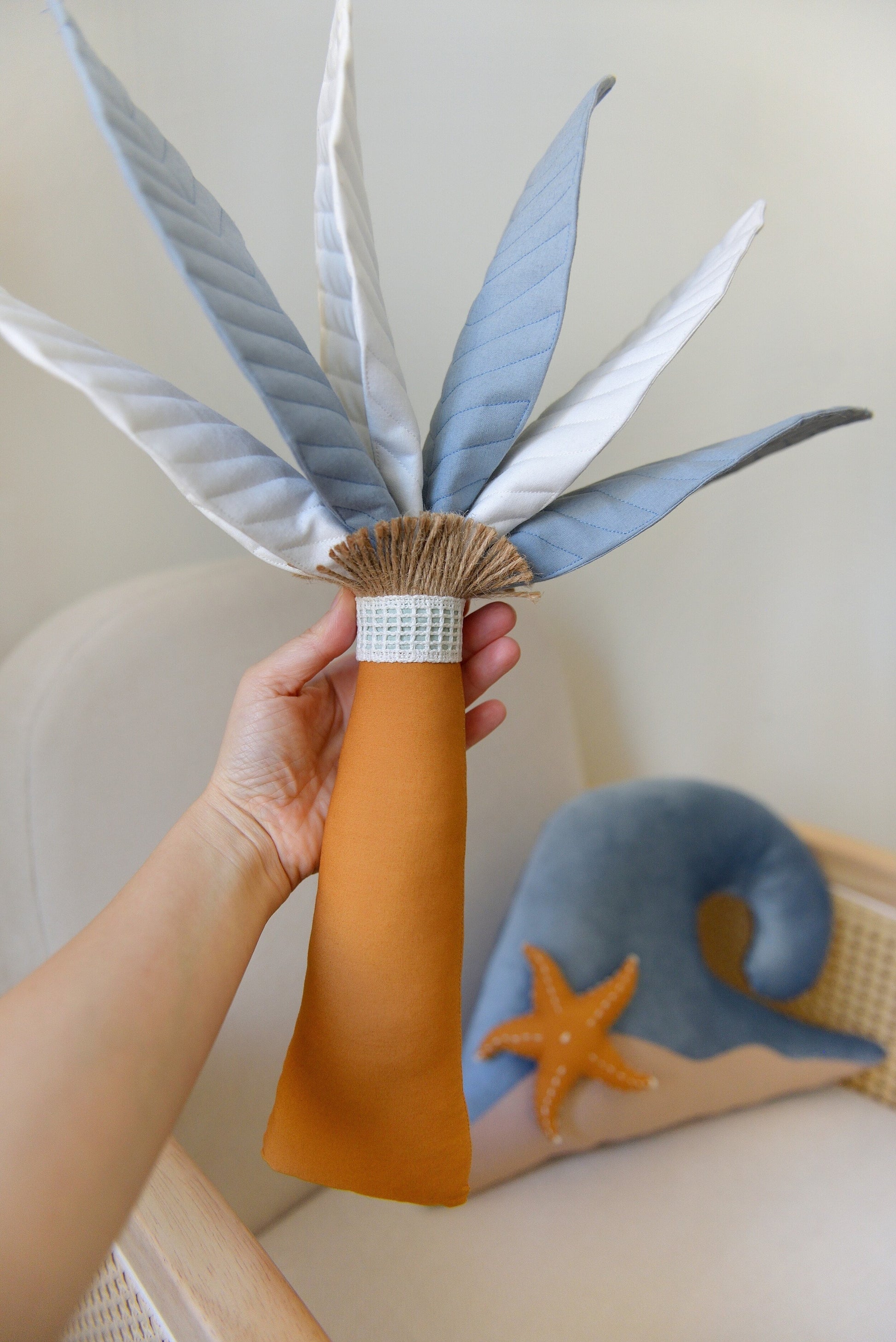 Hand holding Steal Blue Palm Tree Decor Pillow handmade by Boho Baby Heaven. Featuring mixed offwhite and steal blue palm fronds and ochre trunk. Wave Decor Pillow on a rattan armchair blurred in the background.
