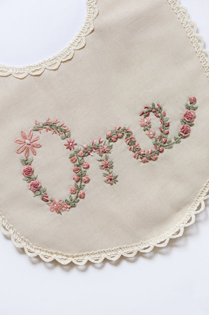 Embroidered floral design spelling ONE on an ivory bib with lace trim