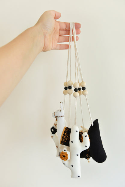 A hand holds a group of handmade boho play gym toys: a black barell cactus play gym toy with rust spikes and ivory flowers, a black crescent moon, two white cacti with rust and black spikes and crocheted flowers and a llama with a rust saddle embellished with vintage style black lace. All hanging of strong macrame rope embellished with 3 wooden beads in the middle.
