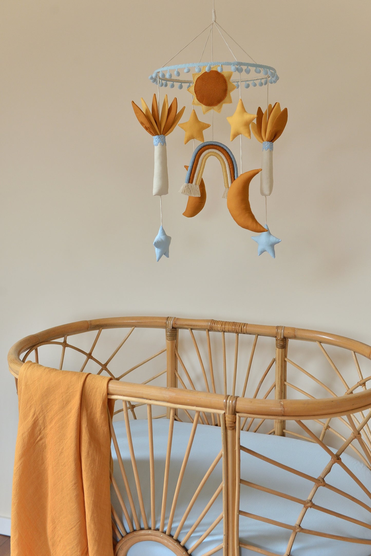 Baby nursery mobile featuring sun, stars, rainbow, and moons and palm trees in ivory, sky blue, yellow and rust shades. It hangs above a rattan bassinet with blue mattress and a swaddle hanging on the left.