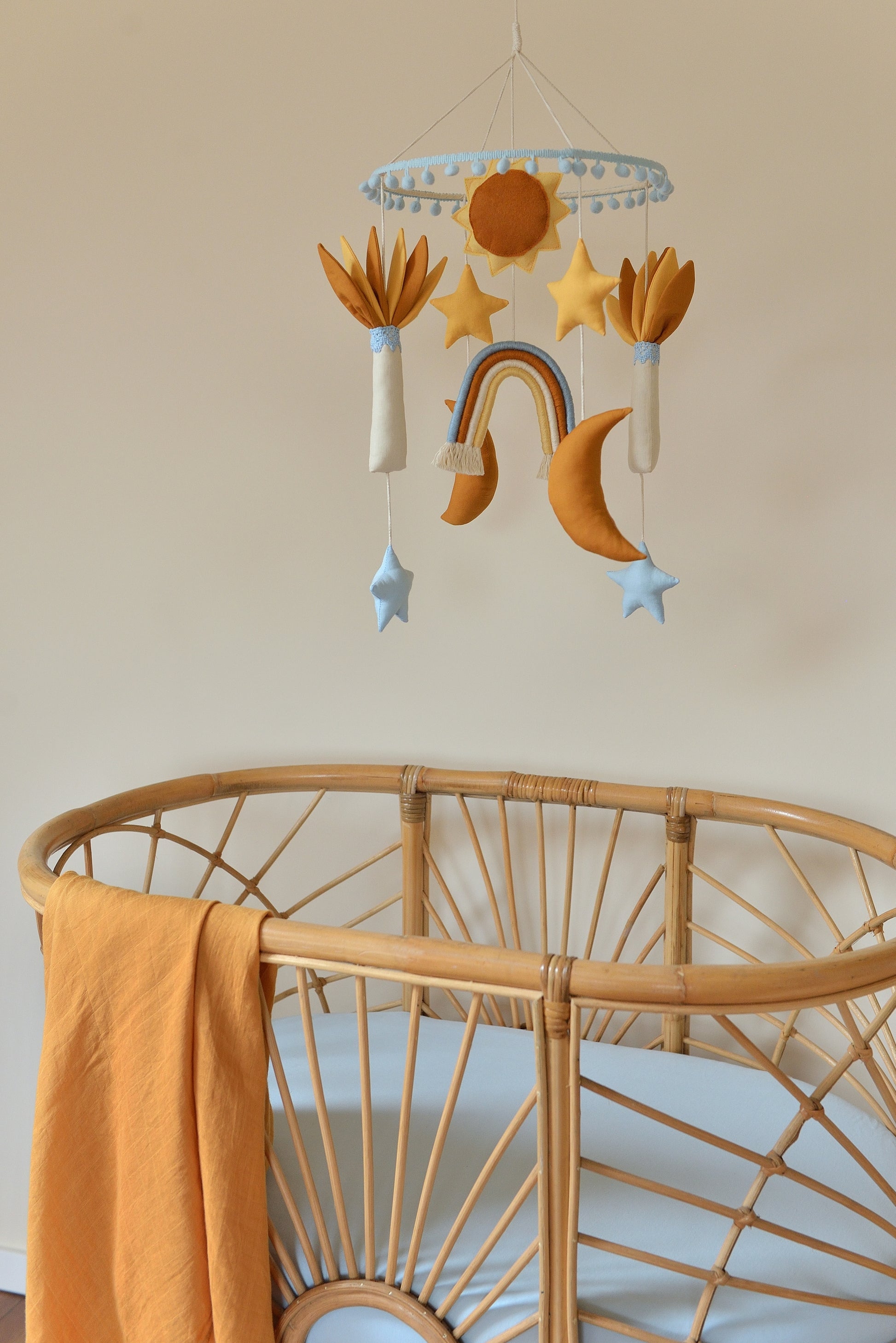 Baby nursery mobile featuring sun, stars, rainbow, and moons and palm trees in ivory, sky blue, yellow and rust shades. It hangs above a rattan bassinet with blue mattress and a swaddle hanging on the left.