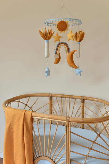 Baby nursery mobile featuring sun, stars, rainbow, and moons and palm trees in ivory, sky blue, yellow and rust shades. It hangs above a rattan bassinet with blue mattress and a swaddle hanging on the left.