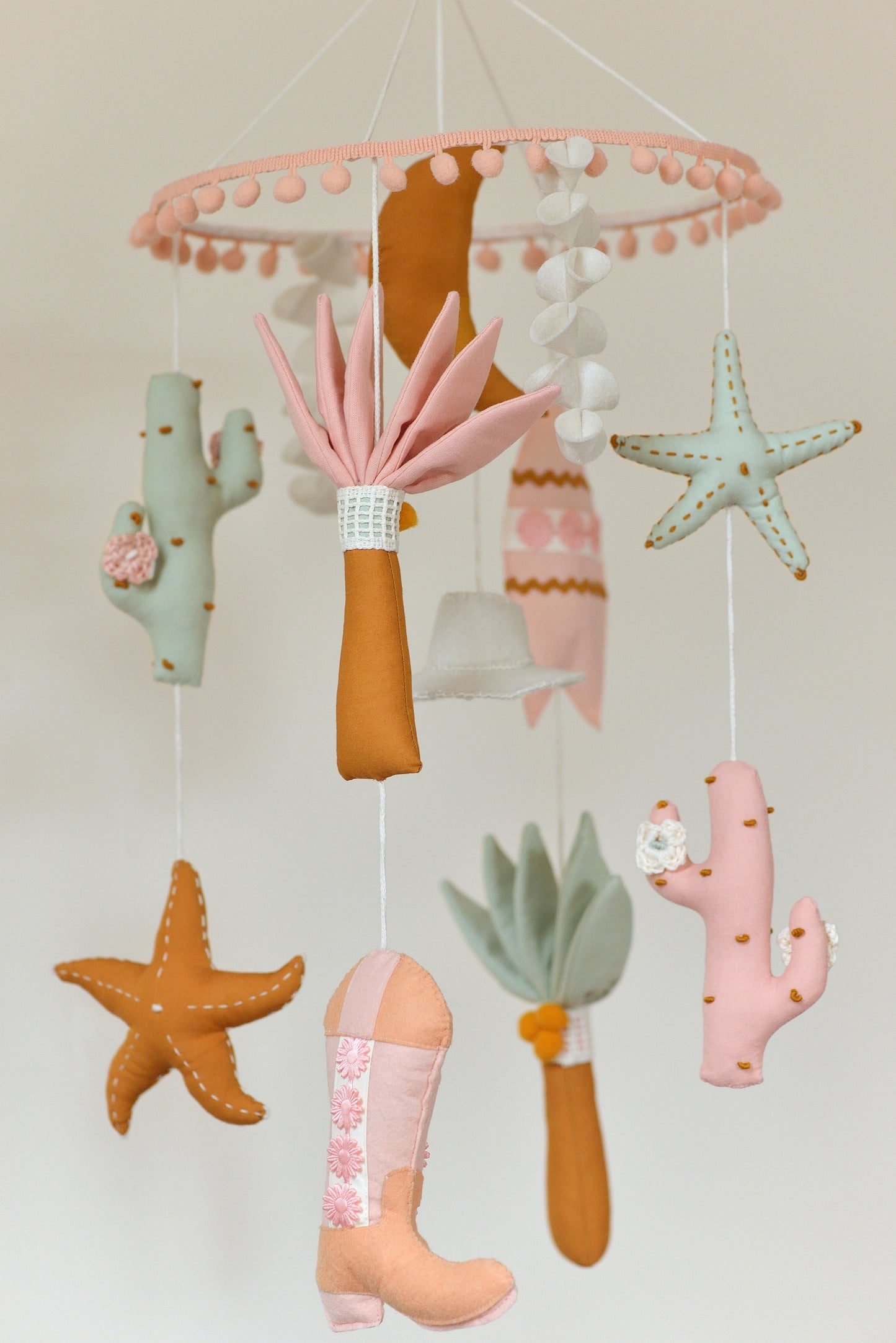 Baby nursery mobile with colorful hanging figures against a light background: moon, palms, starfish, cowboy hat, surf board, cacti, cowboy boots.