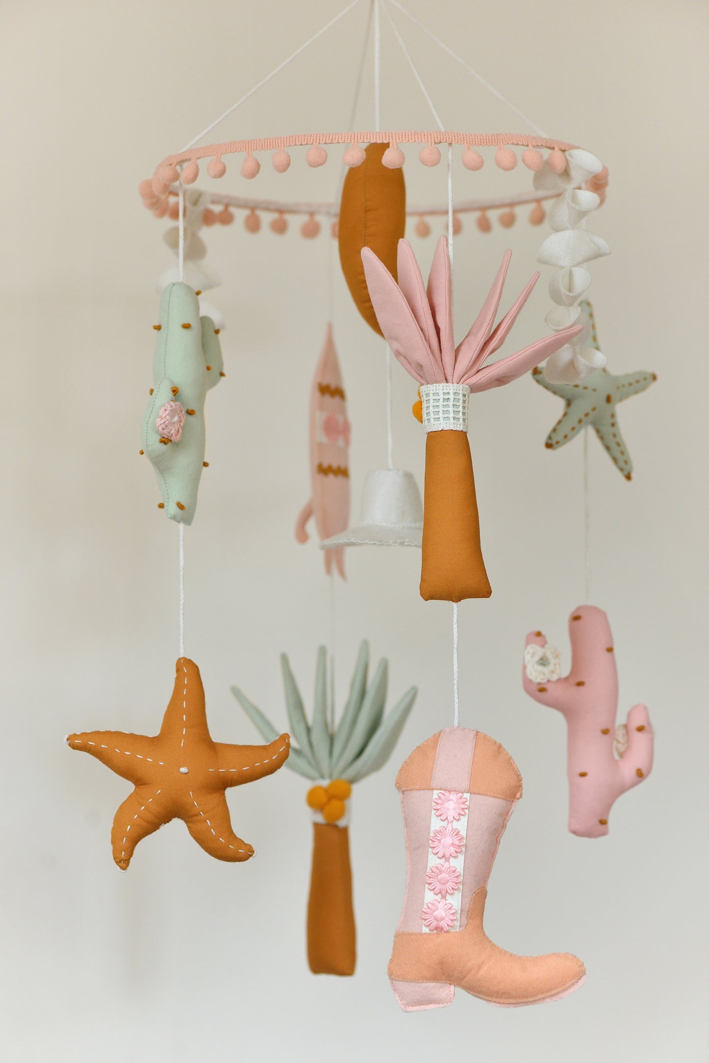 Baby nursery mobile with colorful hanging figures against a light background: moon, palms, starfish, cowboy hat, surf board, cacti, cowboy boots.