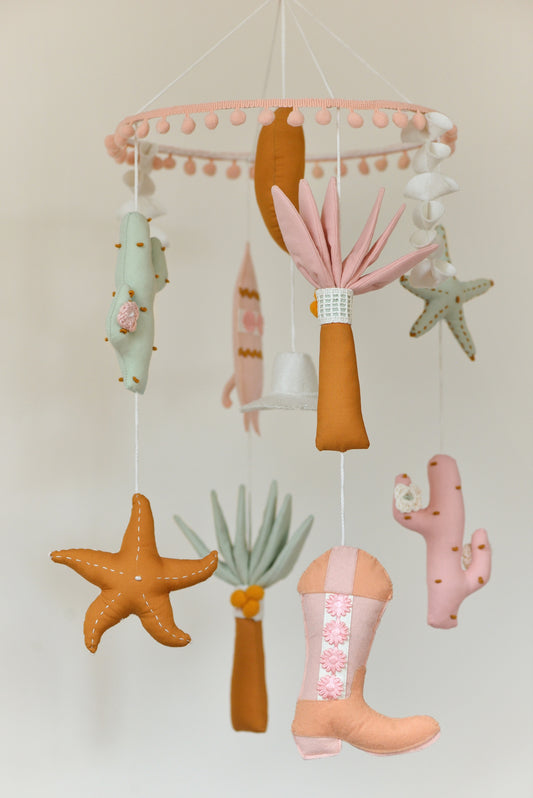 Baby nursery mobile with colorful hanging figures against a light background: moon, palms, starfish, cowboy hat, surf board, cacti, cowboy boots.