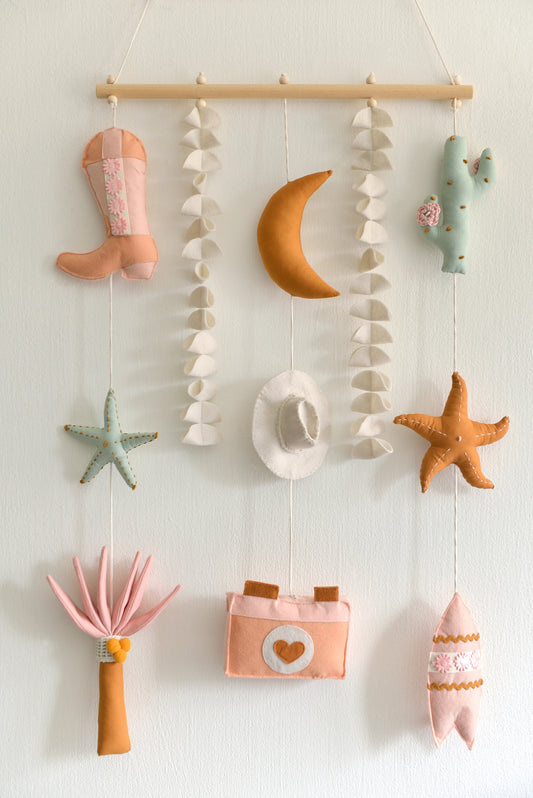 Decorative wall hanging with various coastal figures including a moon, cowboy boot, hat, cactus, starfish, palm and camera against a light wall.
