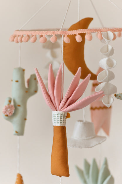 Baby nursery mobile with colorful hanging figures against a light background: moon, palm, cactus, cowboy hat, surf board.