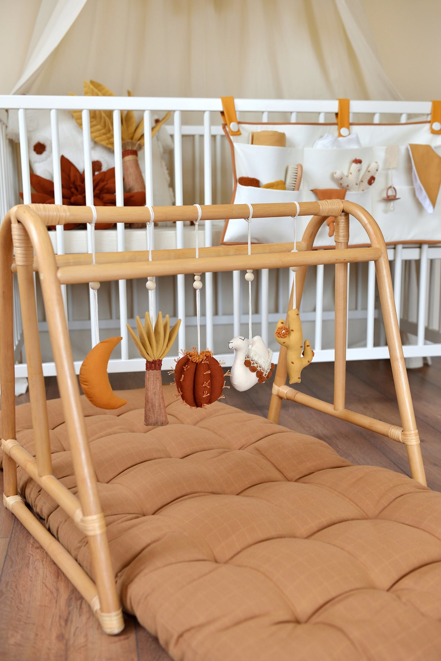 Desert themed earthy colour baby play gym toys hanging on a rattan gym stand: palm, moon, cacti, camel. White crib with hanging crib organizer and baby essentials in the background.