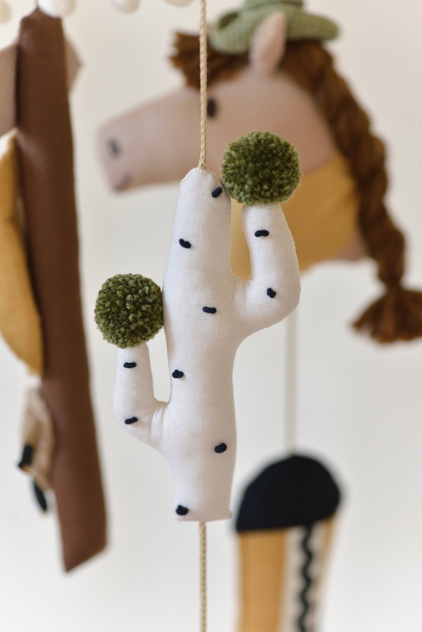 Closeup of handmade Wild West rodeo nursery mobile with cowboy boots, hats, horses, cacti, stars, and rodeo signpost in boho desert style.