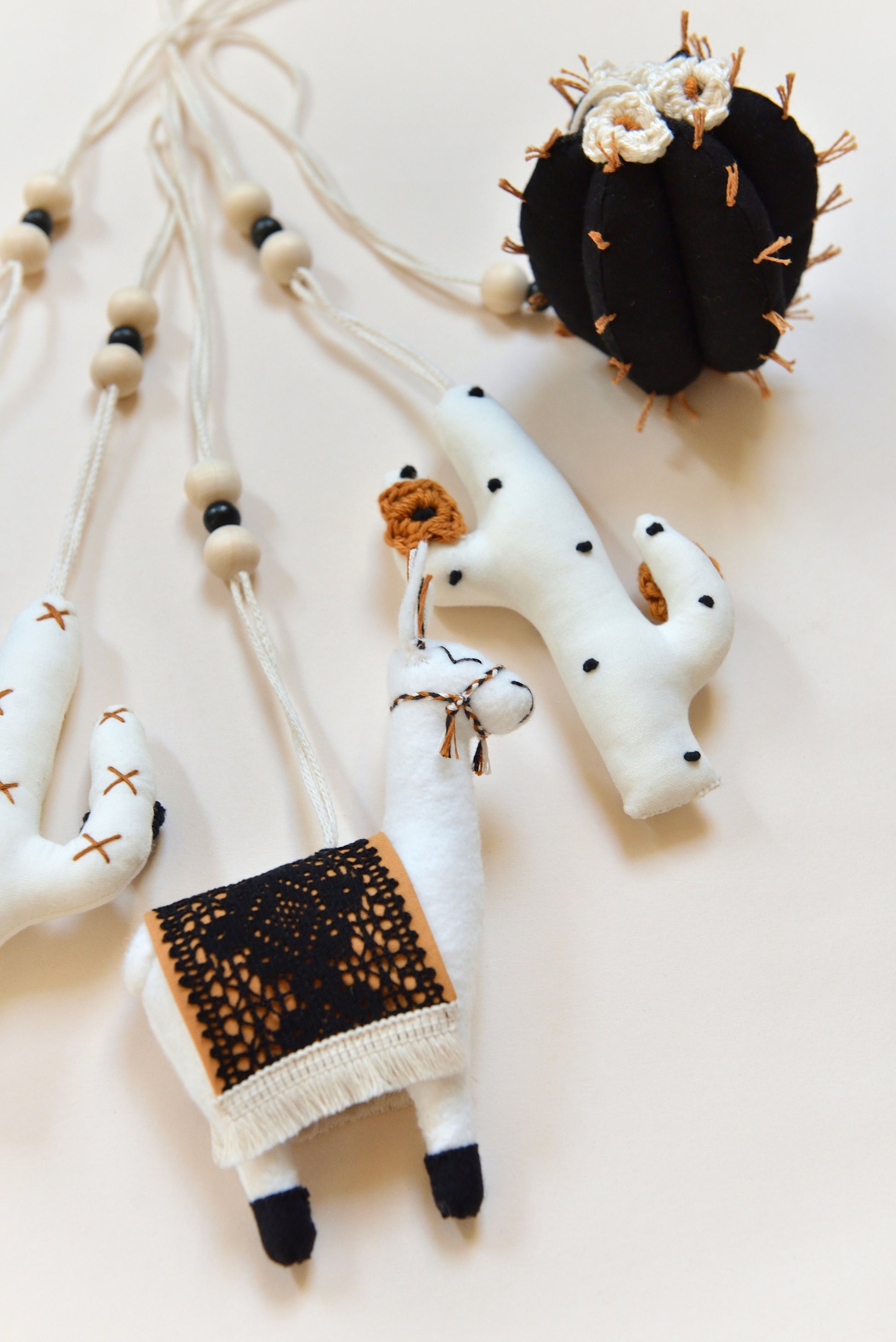 Close up of  flatlay featuring four handmade boho baby play gym toys on an ivory background: two white cacti with rust and black spikes and crocheted flowers, a llama with a rust saddle embellished with vintage style black lace, and a round black barell cactuc with rust spikes and ivory flowers. All hanging of strong macrame rope embellished with 3 wooden beads in the middle.
