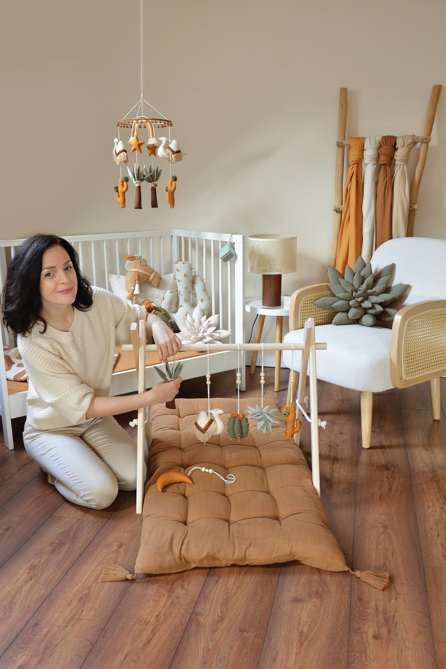 Desert themed baby room with white crib, rattan armchair. Woman attaching hanging toys to play gym in the foreground.