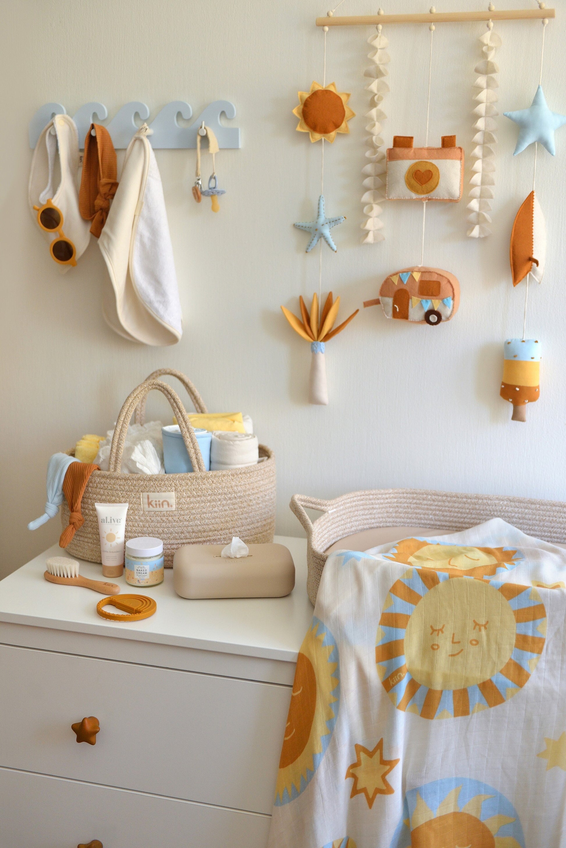 Nappy changing station with baby items  on a white dresser. Surf-themed wall hanging and wave-shaped wall hanger above the dresser. Nappy caddy and nappy changing basket with sun themed swaddle on top of the dresser.