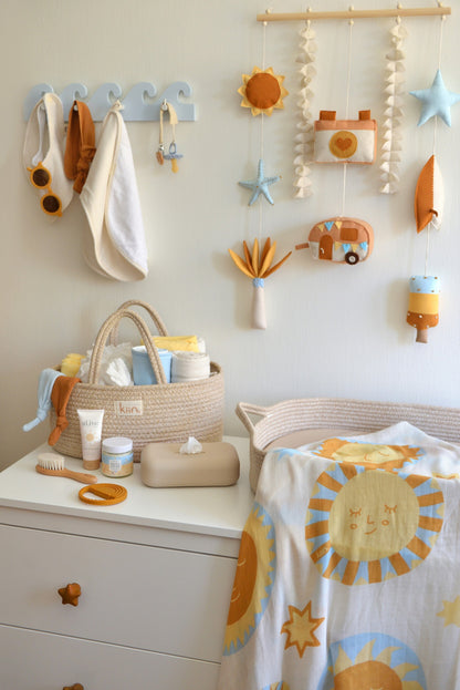 Nappy changing station with baby items  on a white dresser. Surf-themed wall hanging and wave-shaped wall hanger above the dresser. Nappy caddy and nappy changing basket with sun themed swaddle on top of the dresser.