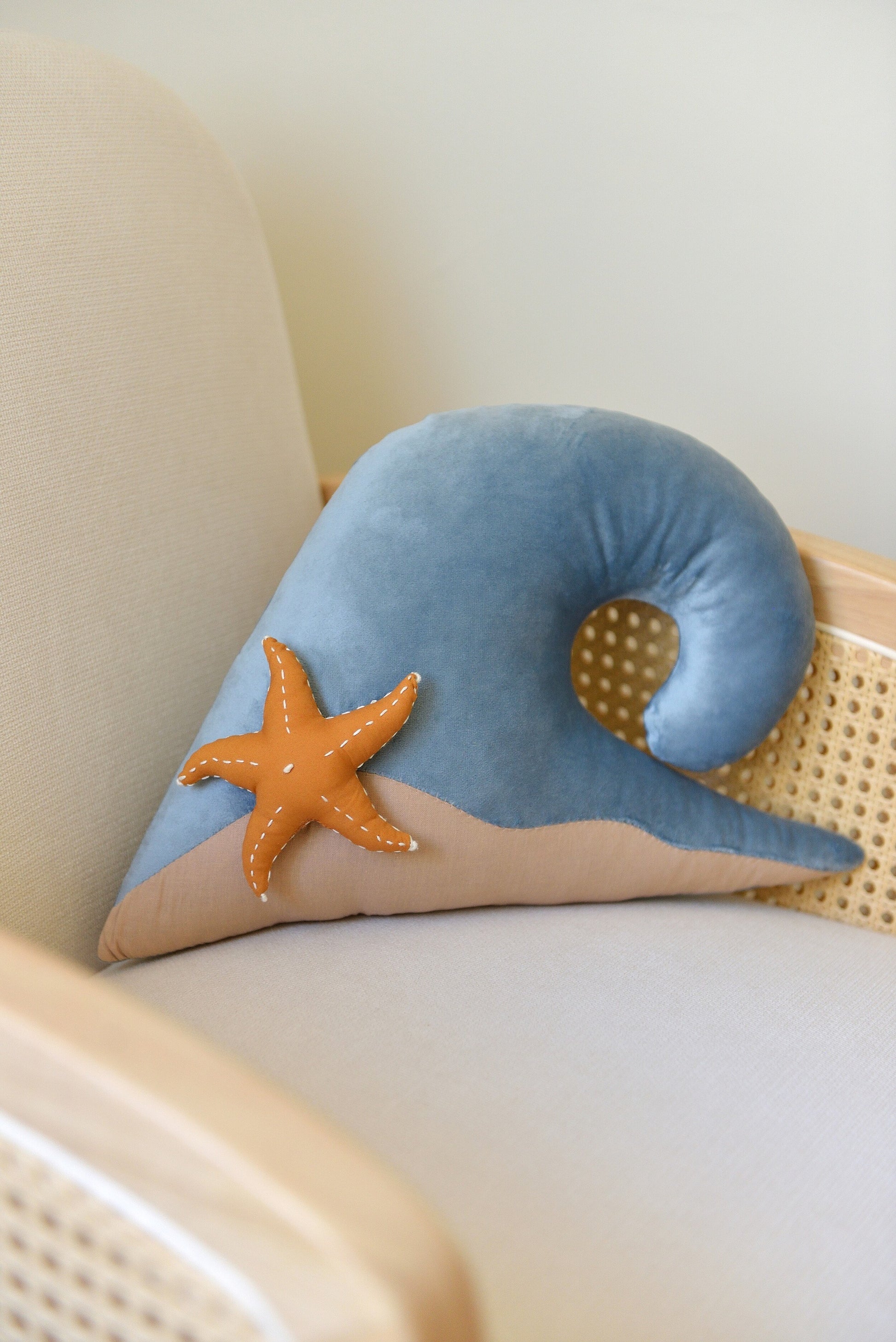Ocean wave shaped decor pillow with a starfish placed on a rattan armchair against a white wall.
