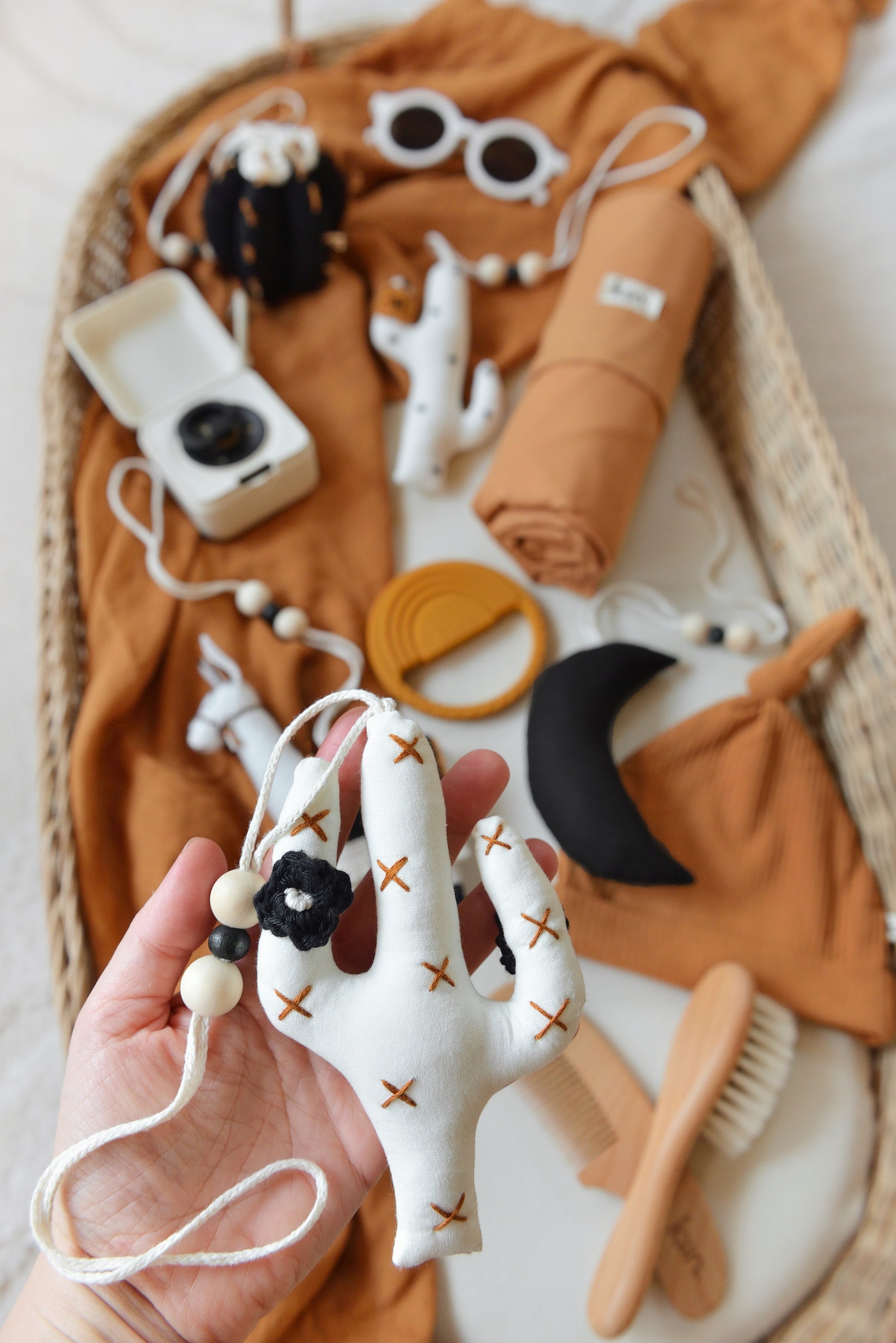 A hand holds an offwhite cactus baby play gym toy. In the background a wicker baby changing basket holds baby items placed on a rust-colored swaddle: sunglasses, five handmade llama, moon and cactus baby play gym toys, pacifier and storage box, cotton beanie, wooden comb set, travel change mat and silicone teether creating a cozy feel.