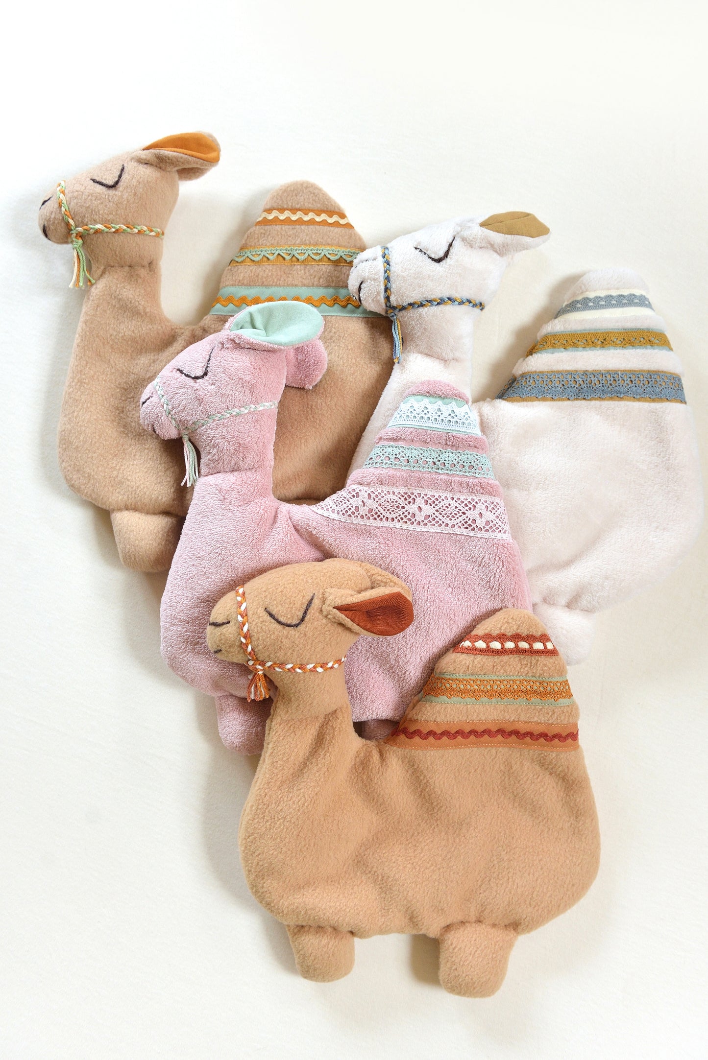 Selection of handmade camel shaped baby comforters with lace details on an ivory background in different colours: ivory, tan and blush pink.