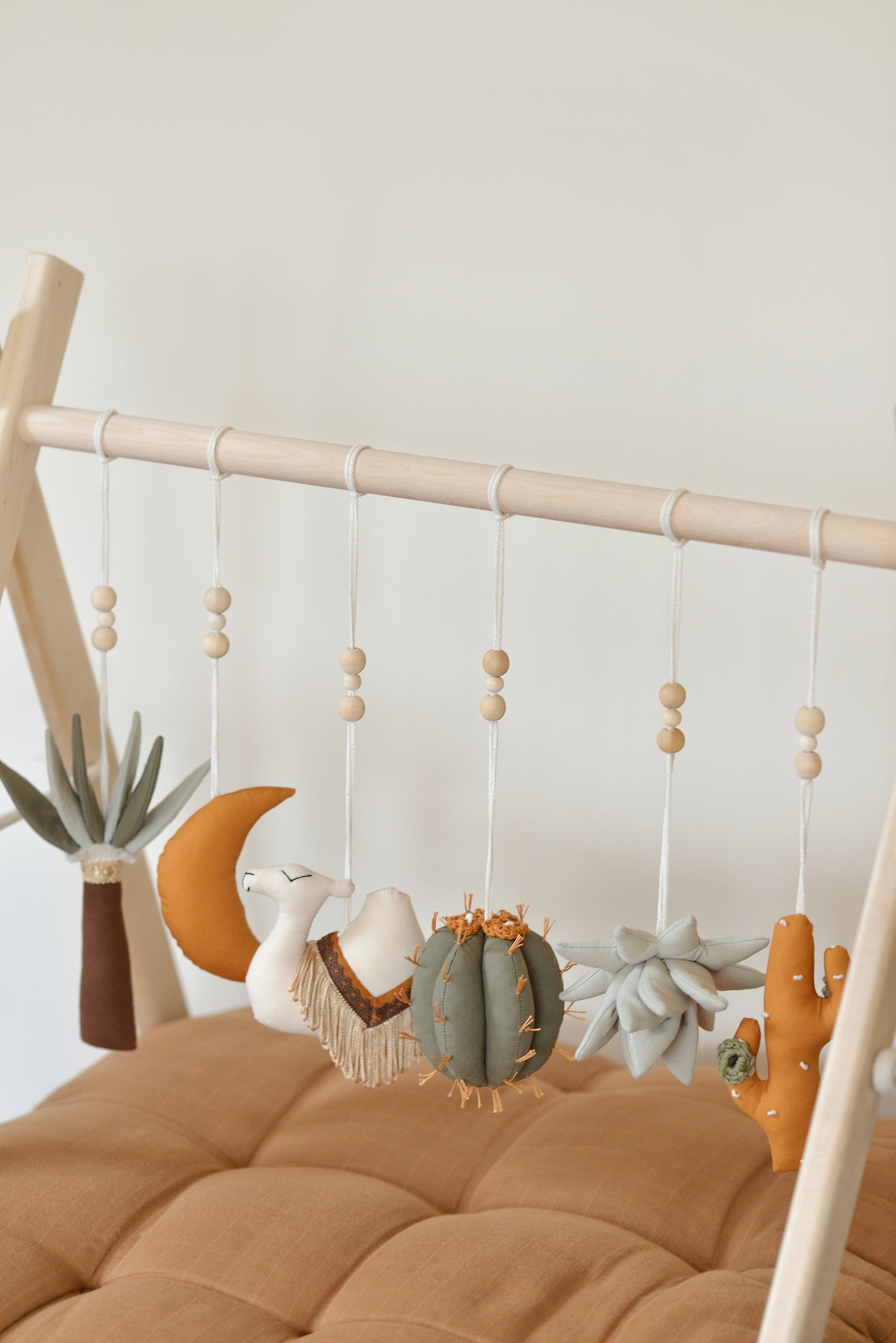 Desert themed play gym toys hanging on a wodden play gym stand: moon, palm, succulent, camel and cacti in khaki, sage, brown and ochre colours. Brown playmat under.