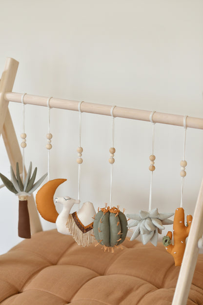 Desert themed play gym toys hanging on a wodden play gym stand: moon, palm, succulent, camel and cacti in khaki, sage, brown and ochre colours. Brown playmat under.