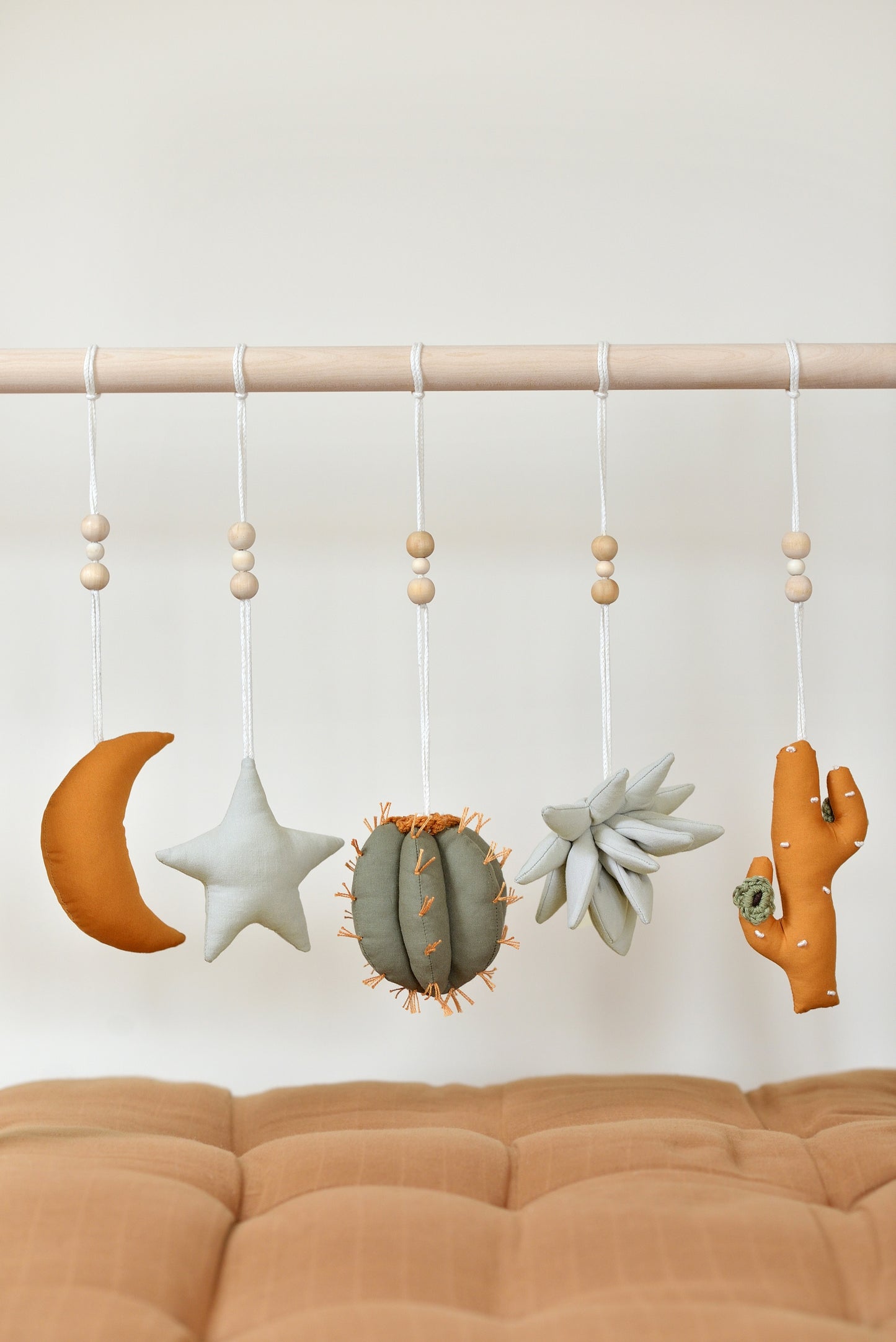 Desert themed play gym toys hanging on a wodden play gym stand: moon, star, succulent and cacti in khaki, sage, brown and ochre colours. Brown playmat under.