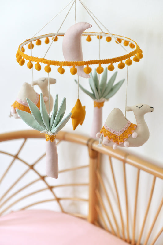 Soft pastel baby nursery mobile with plush shapes hanging over a rattan bassinet with pink sheet under. Featured shapes: ivory camels, mint green palm trees, a marigold yellow star, and a pastel pink crescent moon, creating a whimsical nursery vibe.