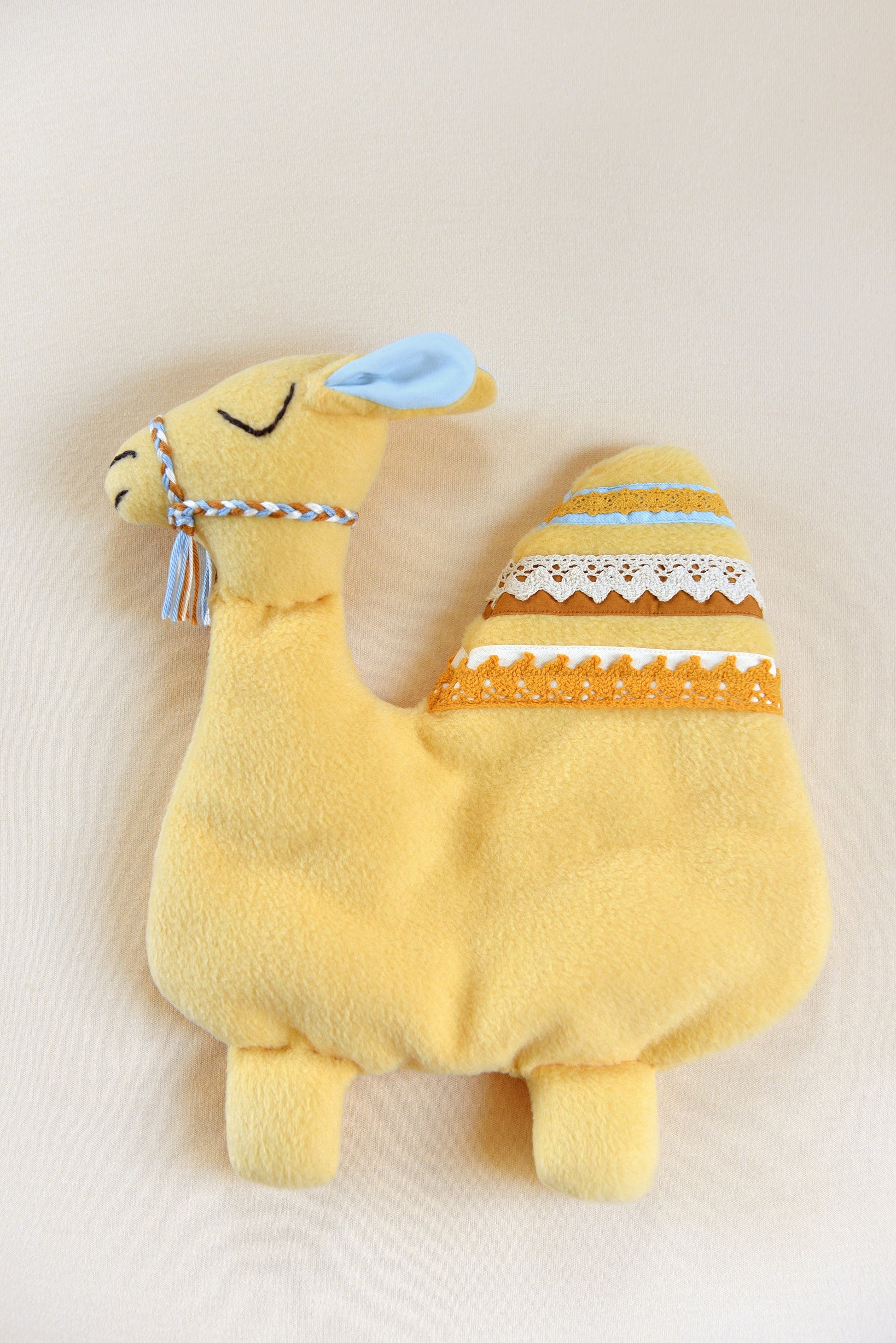 Handmade camel shaped baby comforter with pastel yellow body and white, marigold, blue and rust lace details on an ivory background.