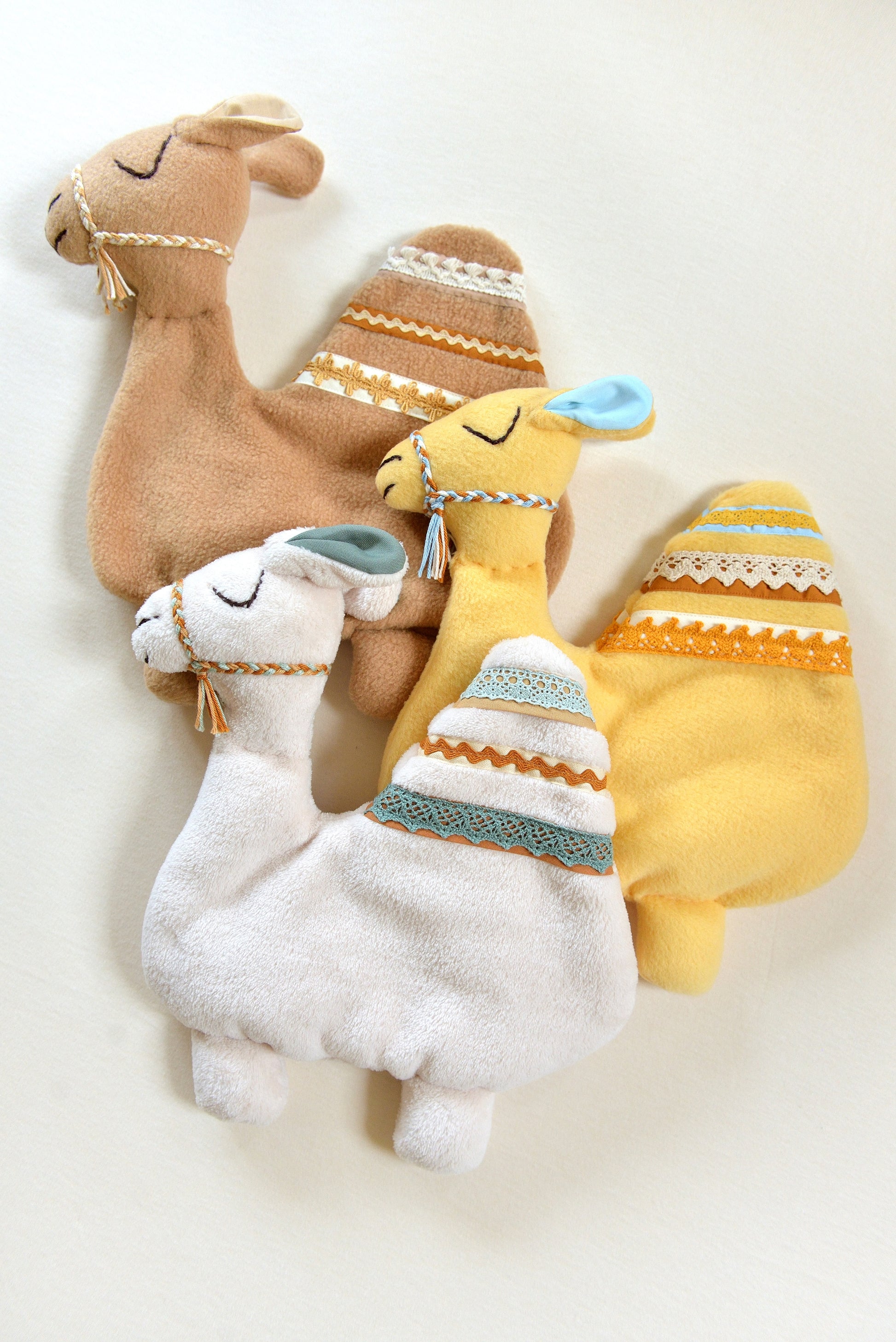 Selection of handmade camel shaped baby comforters with lace details on an ivory background in different colours: ivory, tan and yellow.
