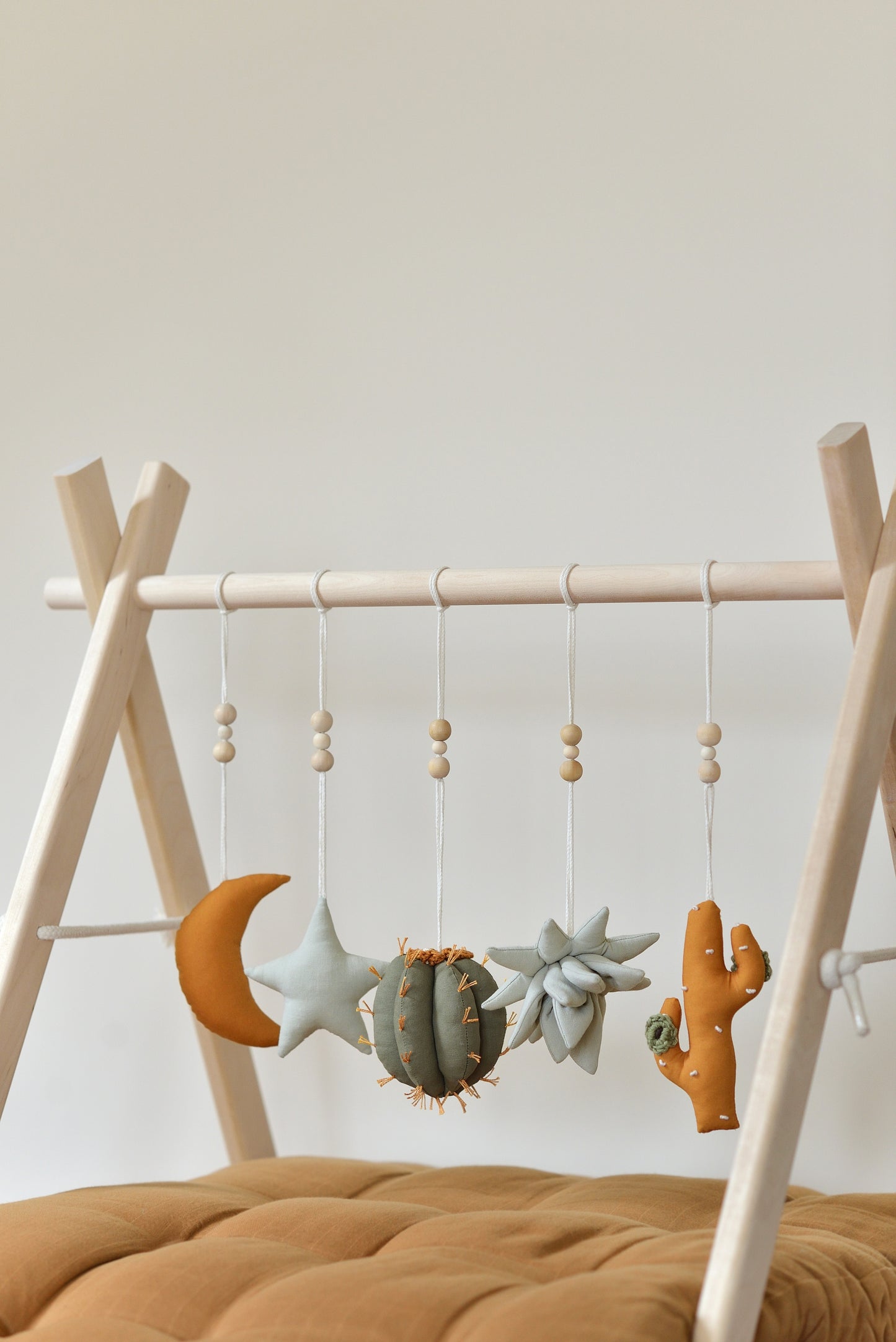 Desert themed play gym toys hanging on a wodden play gym stand: moon, star, succulent and cacti in khaki, sage, brown and ochre colours. Brown playmat under.